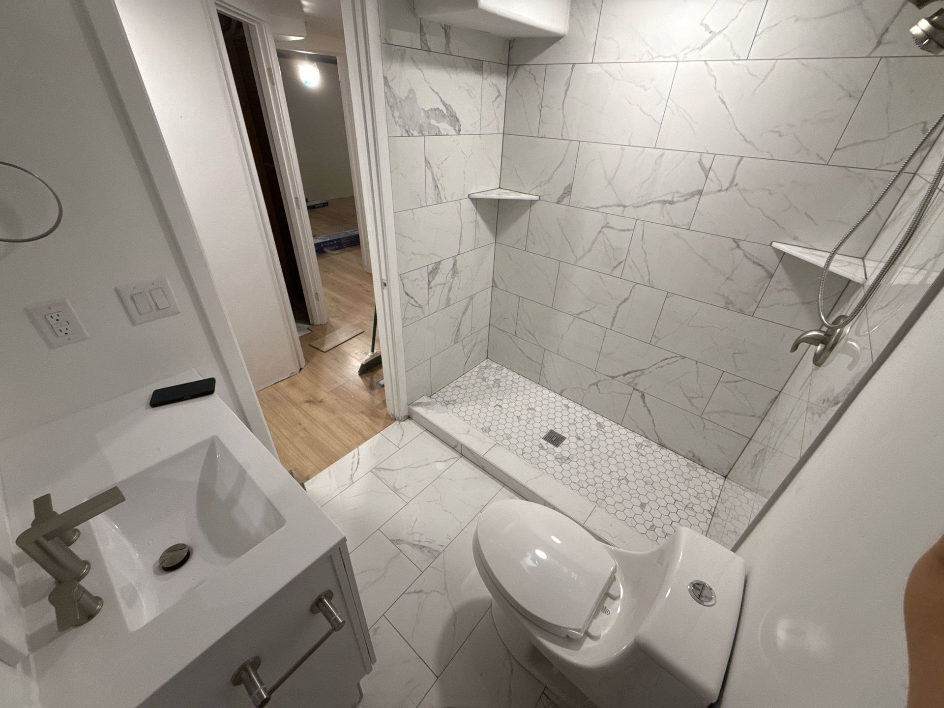 Modern bathroom with white marble tile shower, toilet, and vanity. Hardwood floor visible in the doorway.