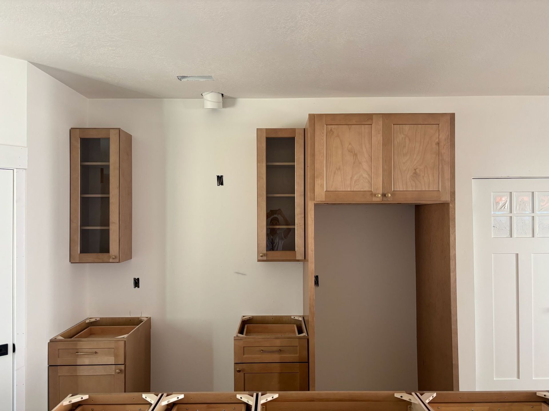 Kitchen renovation: Cabinets in various stages of installation; unfinished wood, some with glass doors, against white walls.