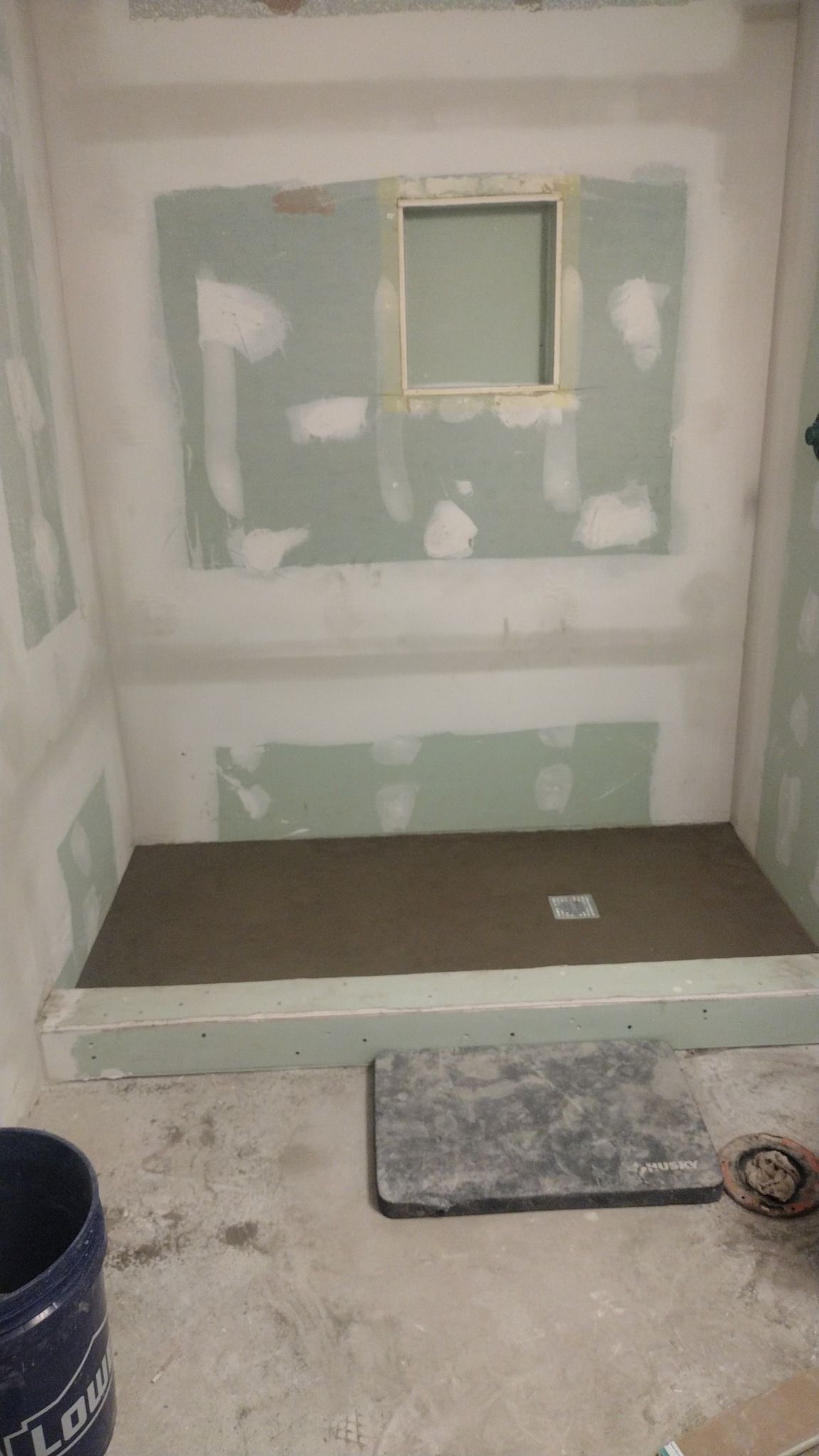 A shower in progress; cement floor, green walls, framed window. Bucket and tile present.
