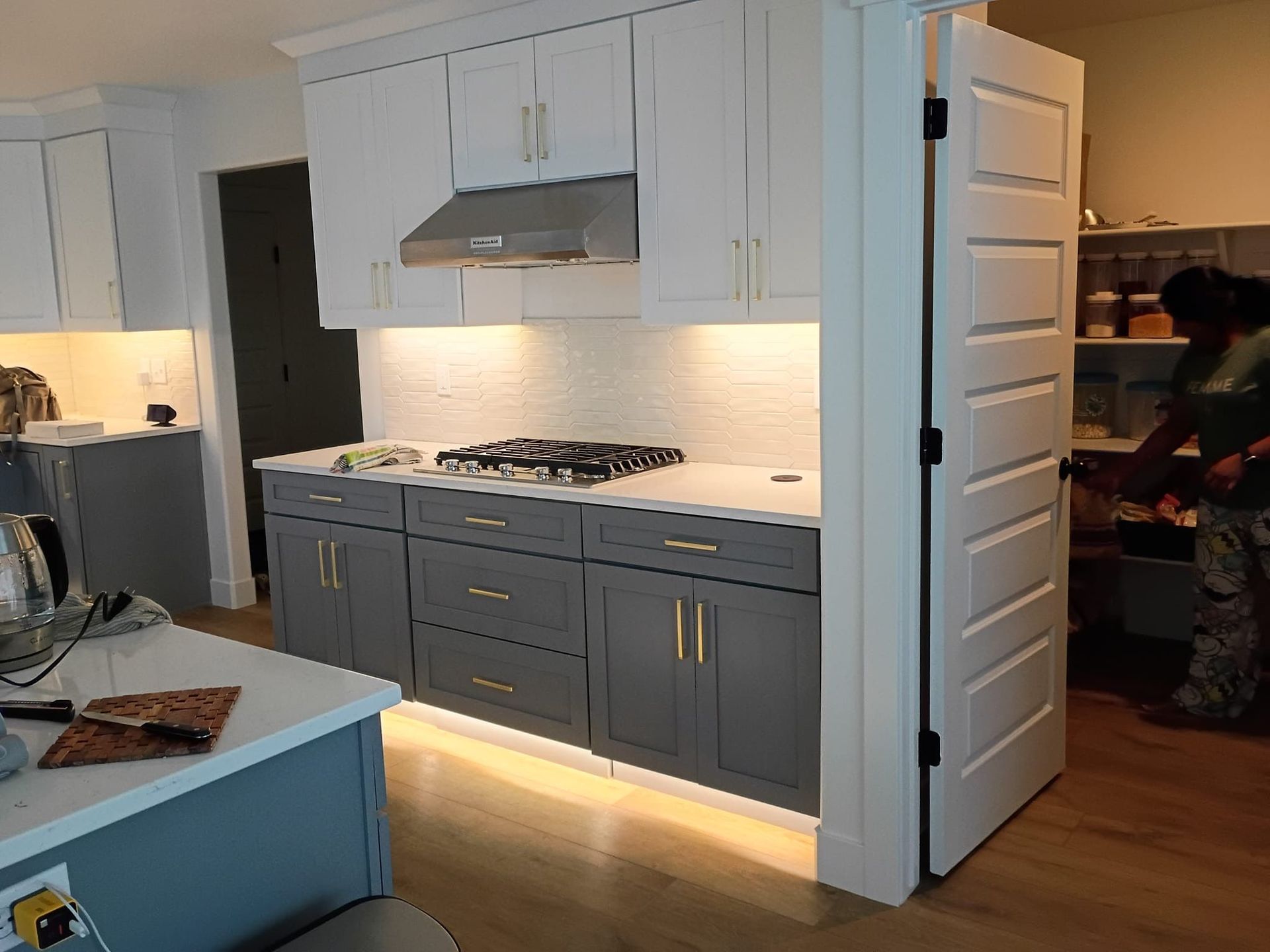 Kitchen with gray and white cabinets, range hood, and lighting. A person reaches into a pantry.