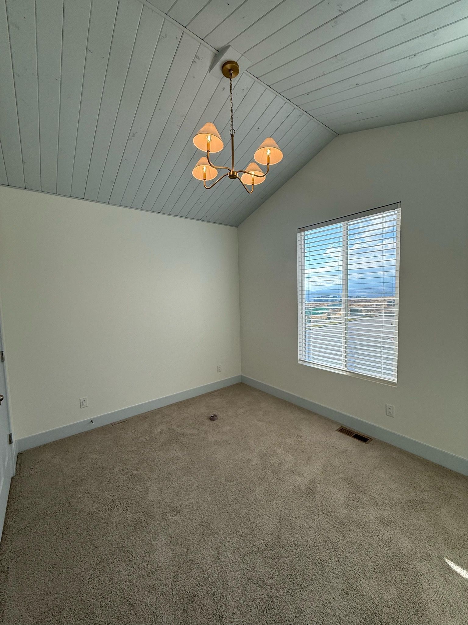 Empty room with vaulted ceiling, window with blinds, beige carpet, and a light fixture.