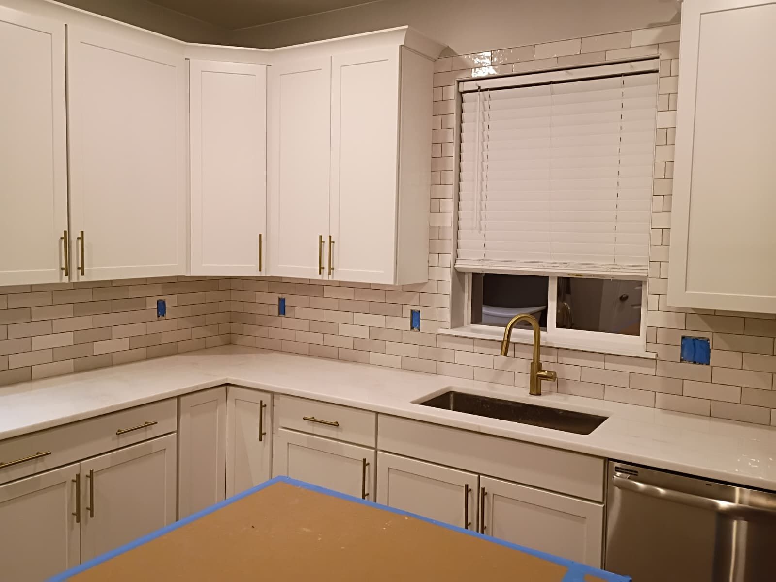 White kitchen cabinets with gold hardware and a mosaic backsplash. Sink and window with blinds.