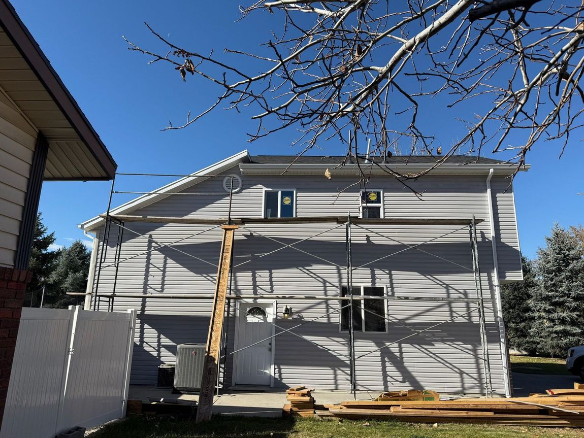 Two-story house with gray siding under construction, scaffolding, lumber, and a clear blue sky.