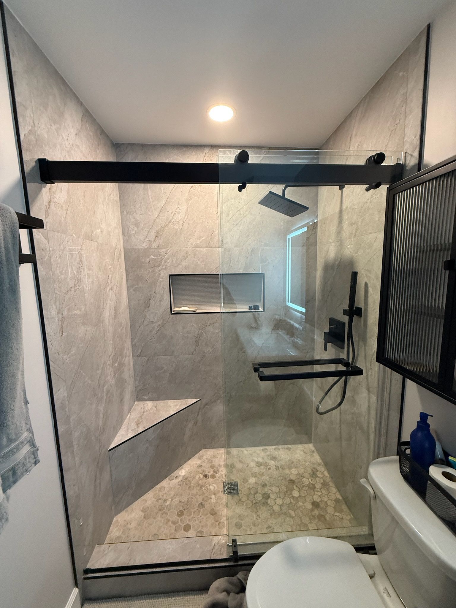 Modern gray-tiled shower with a glass door, black fixtures, and a recessed shelf. A toilet sits nearby.
