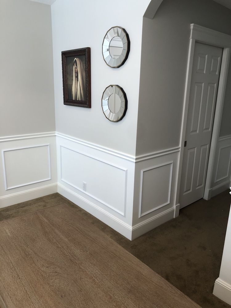 Interior corner with white wainscoting, gray walls, framed artwork, two decorative mirrors, and a closed door.