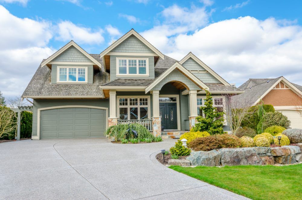 A two-story green-sided suburban home with a gray shingled roof, a paved driveway, and a tiered rock garden out front.