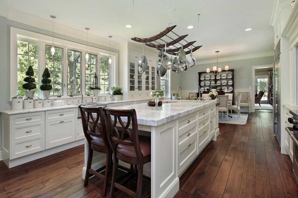 A white kitchen island with bar stools, marble countertops, and a hanging pot rack, set in a room with wood floors.