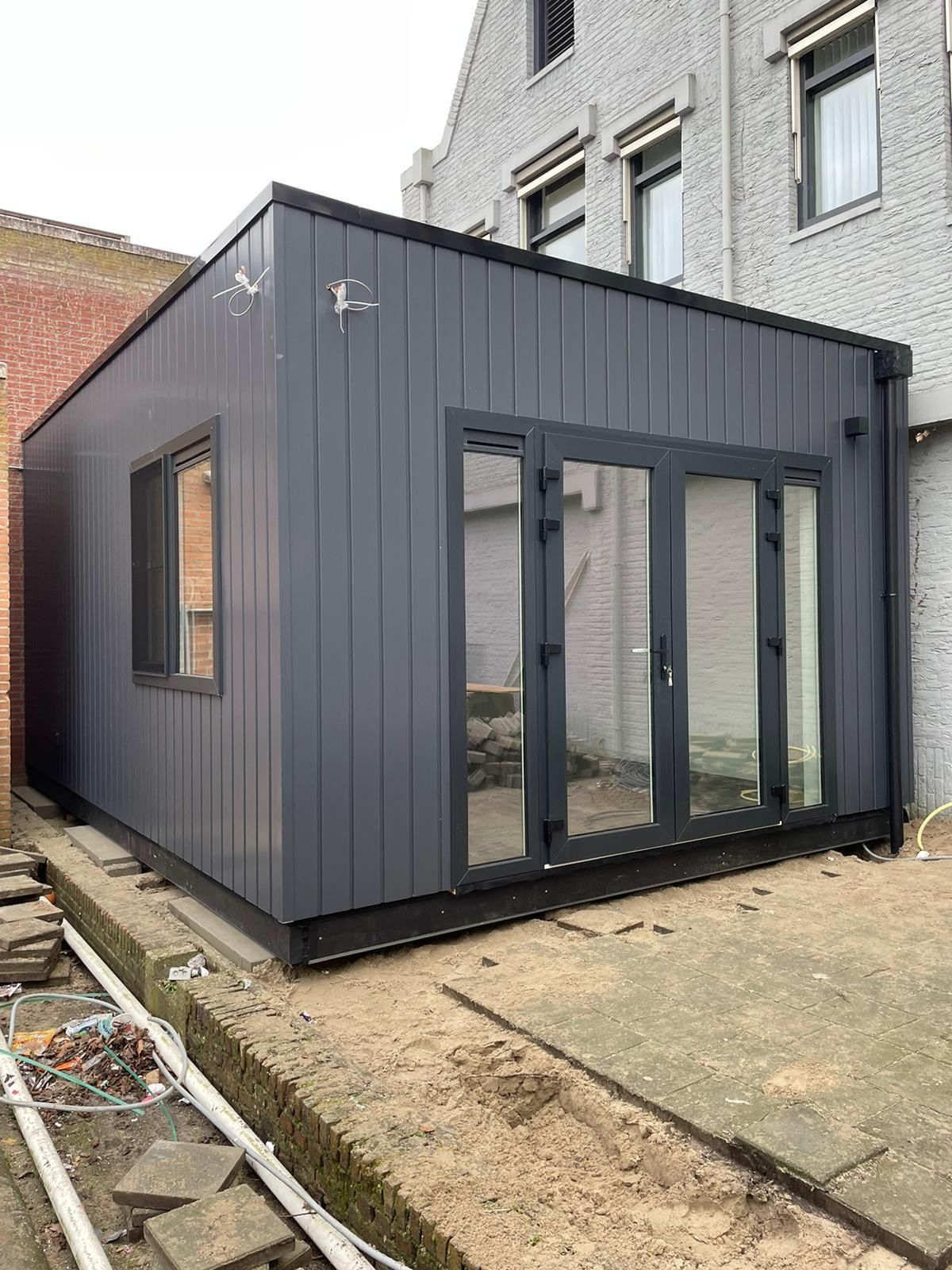 In de achtertuin van een huis wordt een klein gebouw met veel ramen en deuren gebouwd.