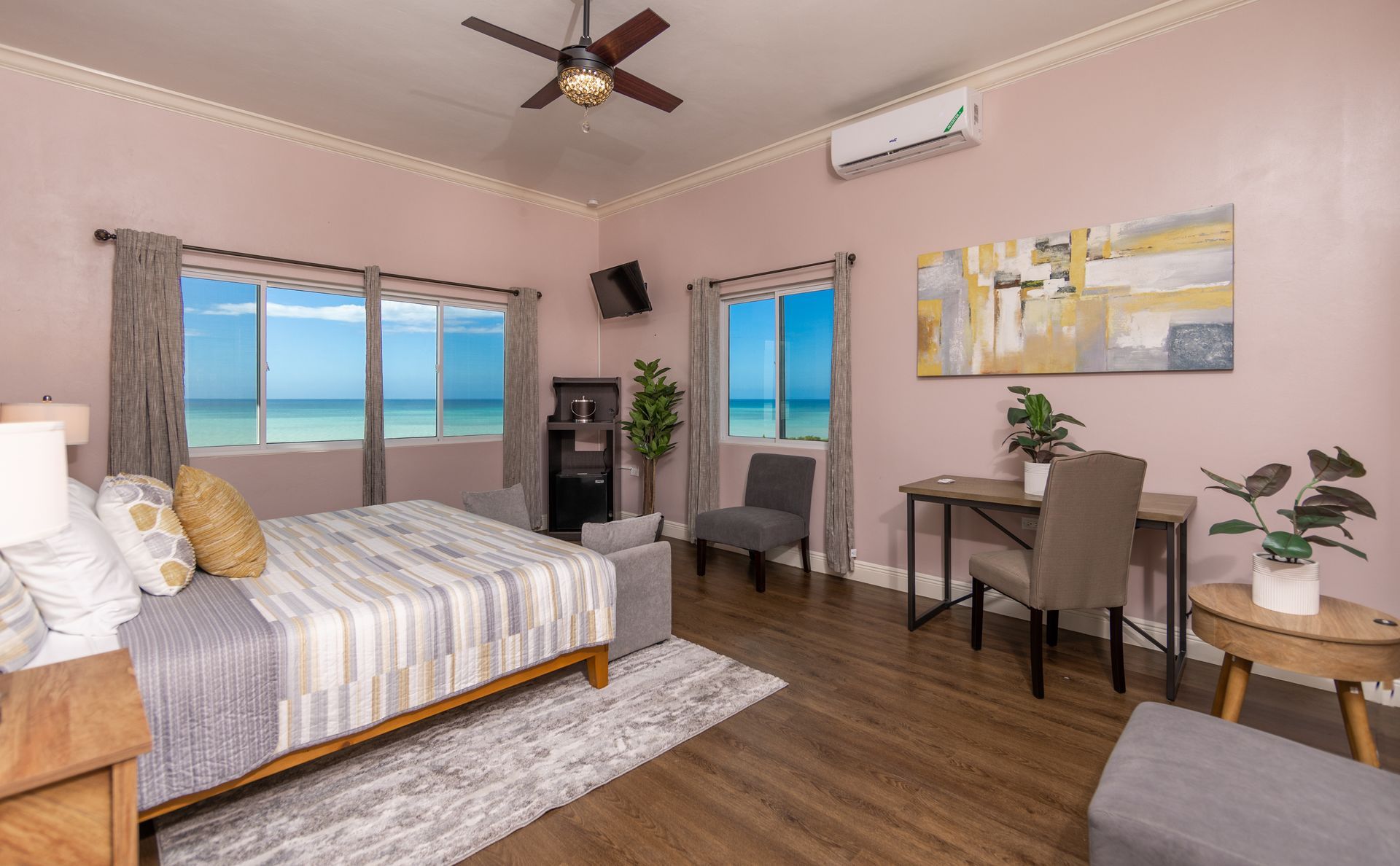 Bedroom with ocean view; bed, desk, art, windows, and plants against a pink wall.