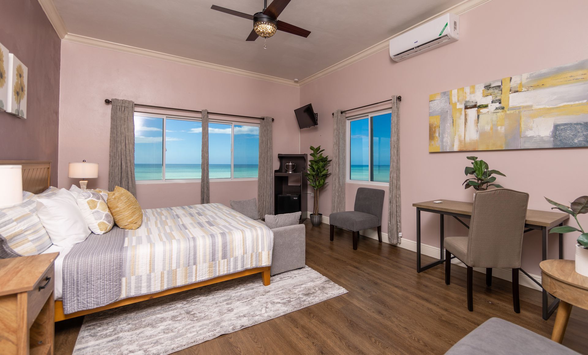 Bedroom with bed, desk, chairs, and large windows overlooking the ocean. Pink walls, brown floor.