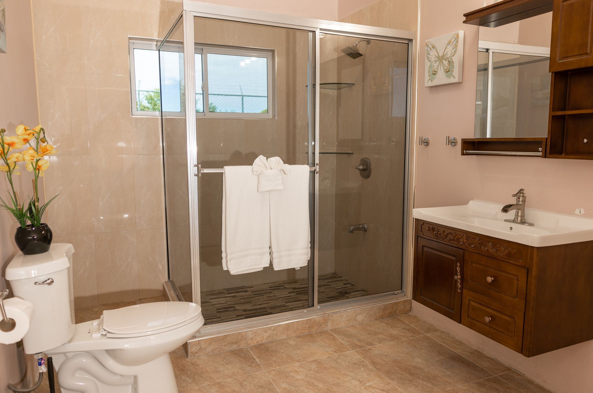 Bathroom with glass shower, toilet, wooden vanity, and beige tile. White towels hang in the shower.