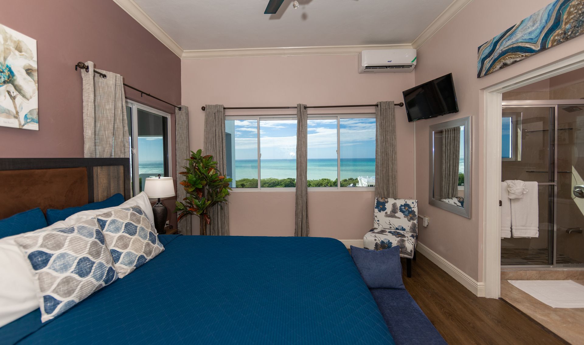 Bedroom with ocean view; blue bedding, artwork, TV, and a bathroom.