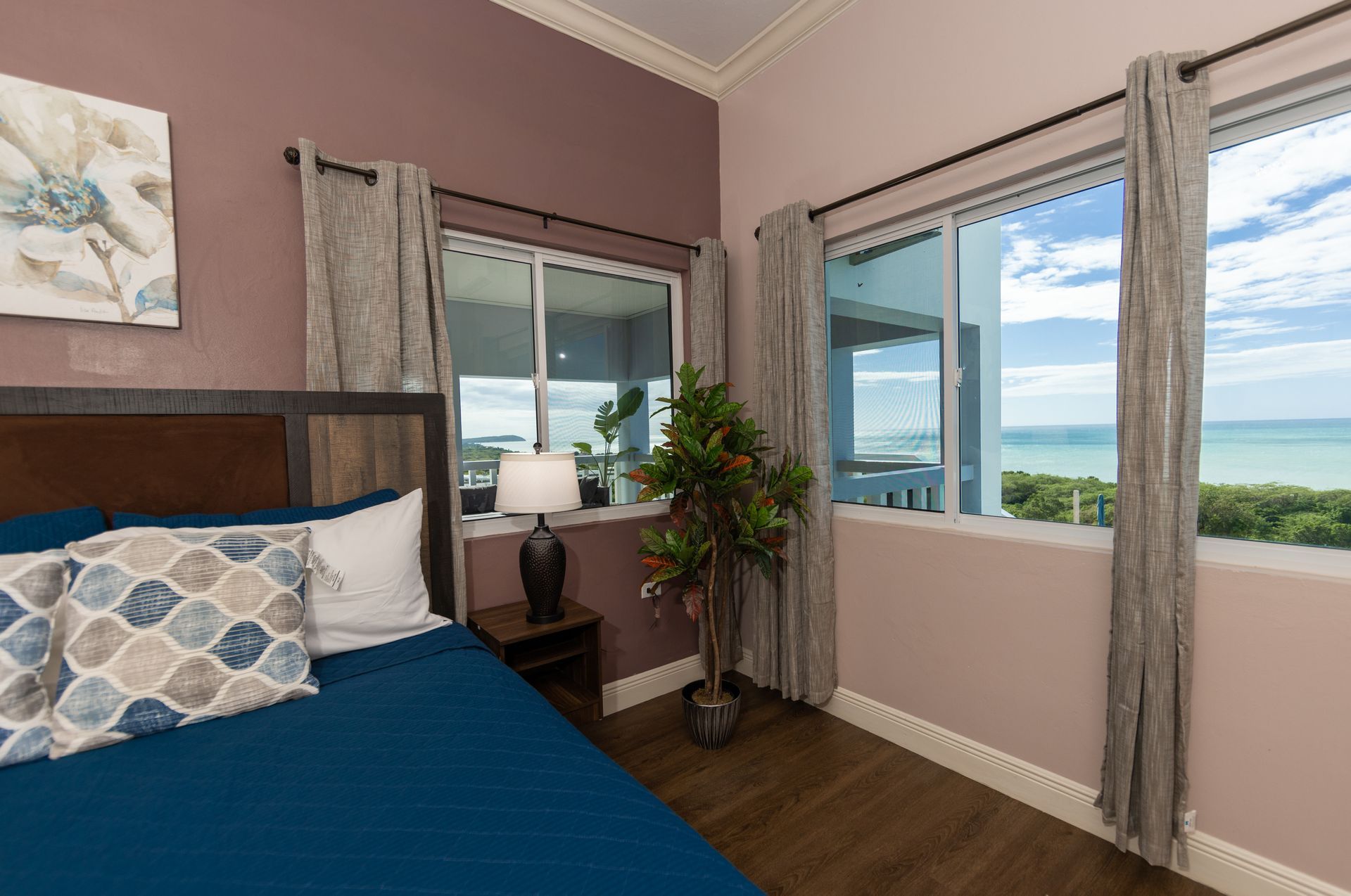 Bedroom with bed, windows overlooking water, art, and plant.