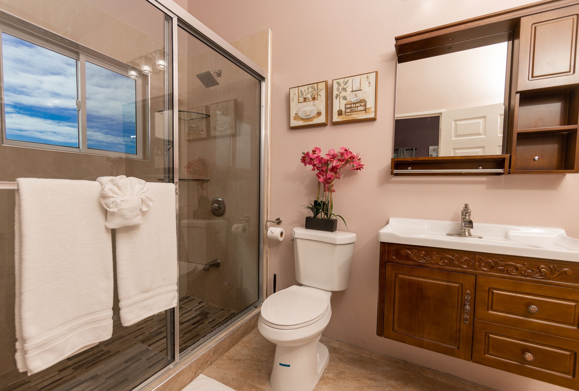 Pink bathroom with shower, toilet, and vanity. White towels hang near the window.