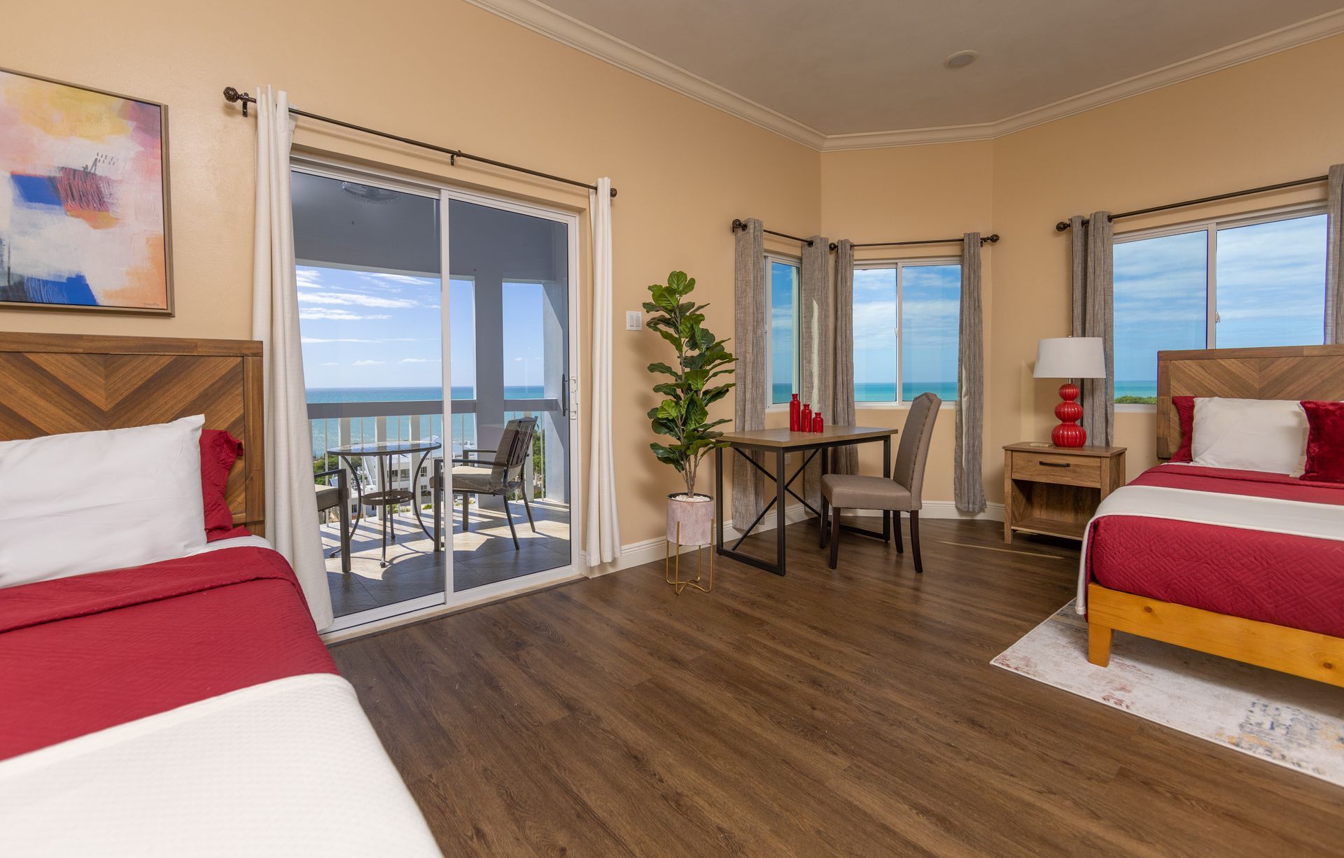 Bedroom with two beds, balcony overlooking the ocean, desk by window, warm tones.