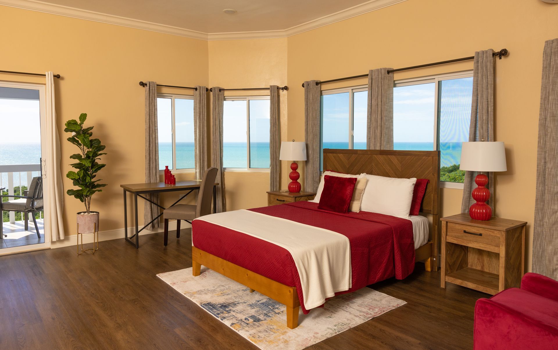 Bedroom with a bed, desk, and windows overlooking the ocean; red and cream bedding, wooden furniture.