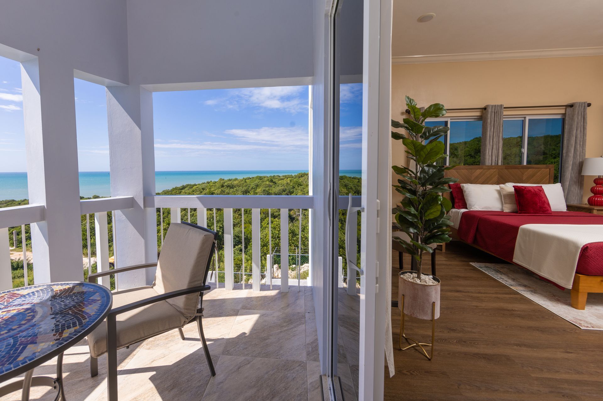 Balcony with ocean view and bedroom interior, door open. Chairs on balcony, bed with red accents inside.