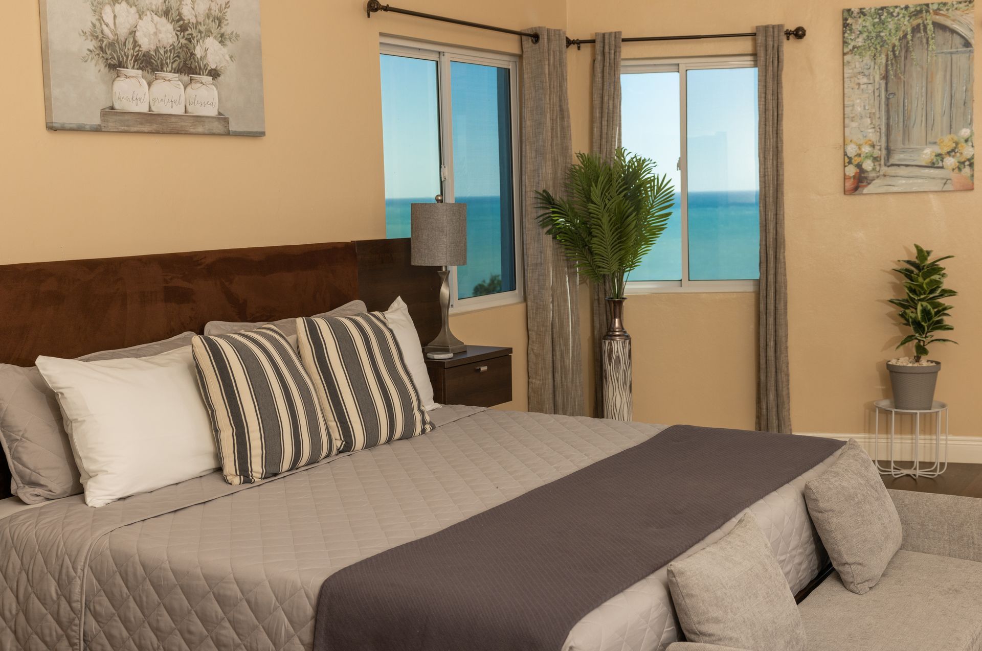 Bedroom with bed, pillows, gray comforter, and a view of the ocean through windows.