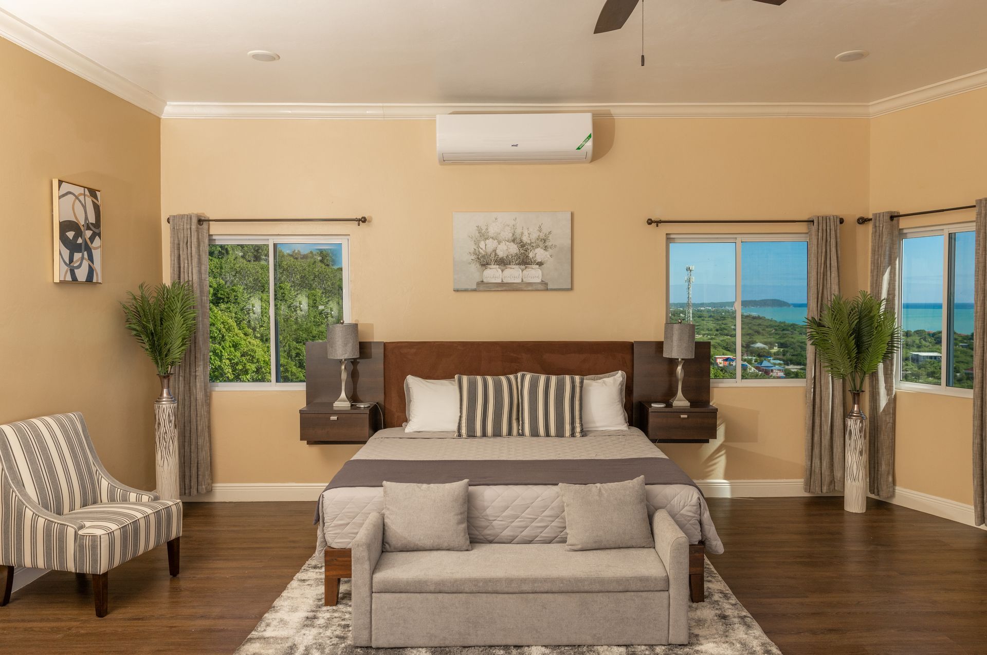 Bedroom with a bed, small sofa, two windows, and chair. Soft colors and a view outside.