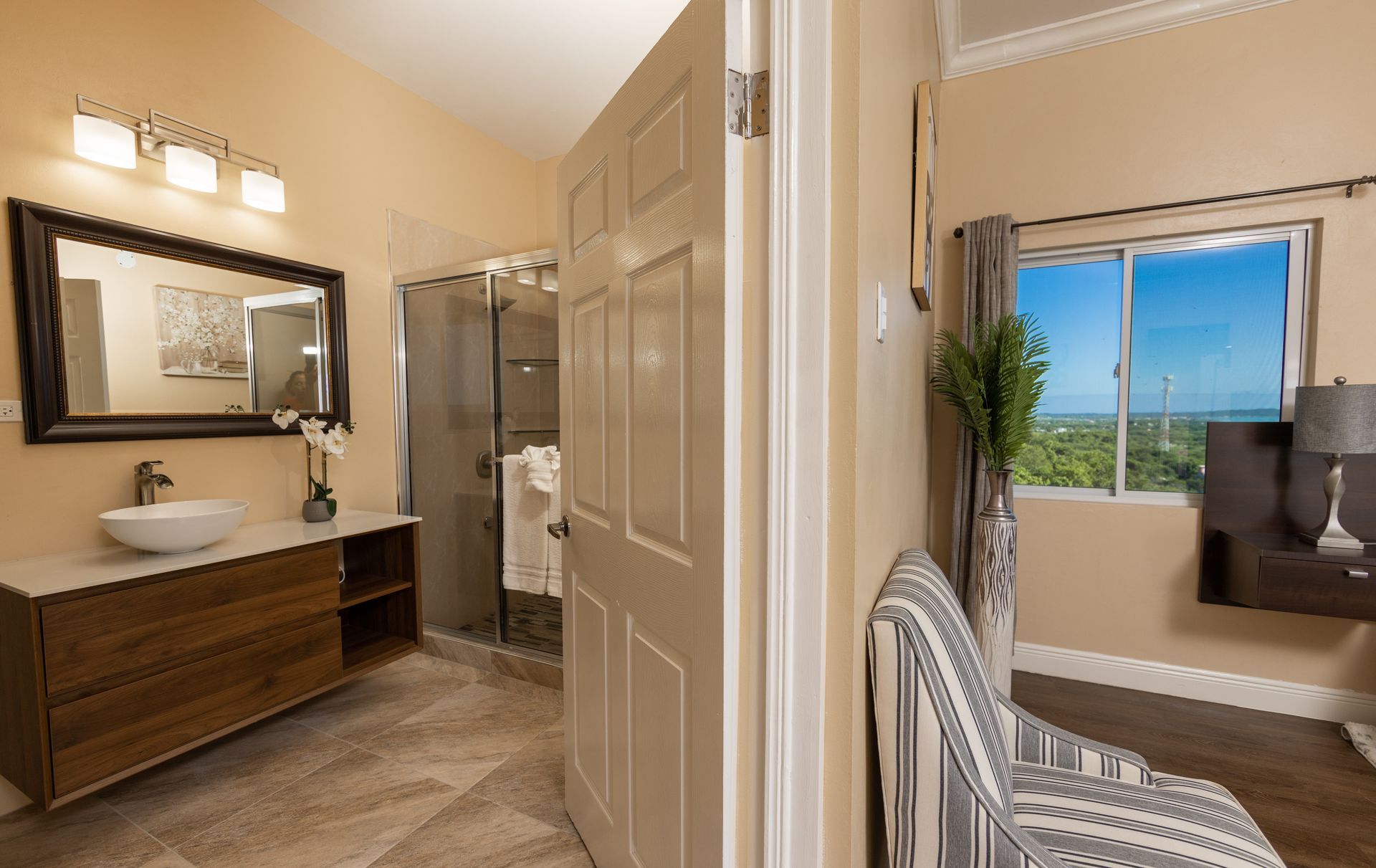 Bathroom doorway open to view a bedroom. Beige walls, wood vanity with sink, glass shower. Window with blue sky.