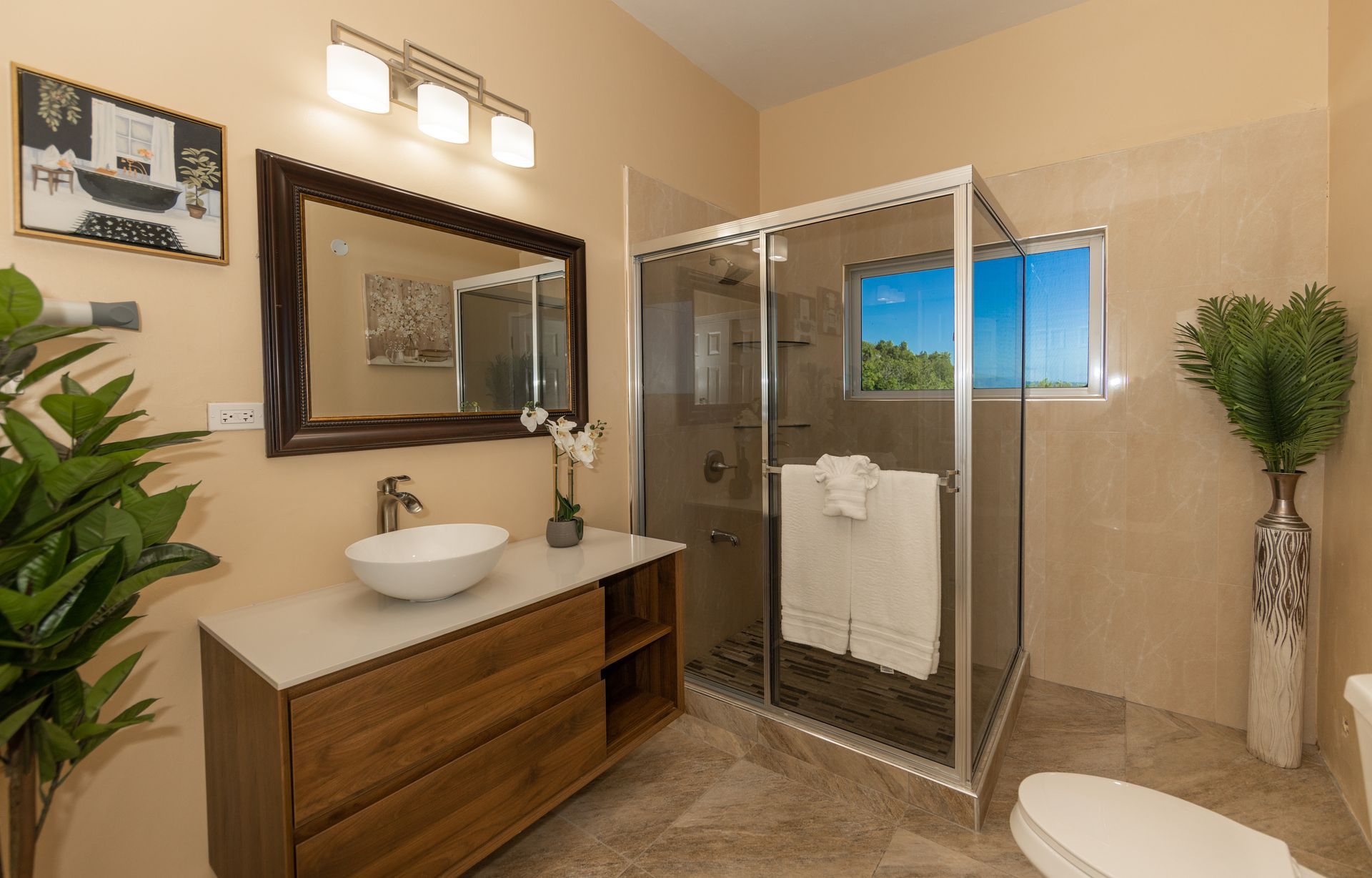 Bathroom with wood vanity, glass shower, large mirror, and decorative plants.