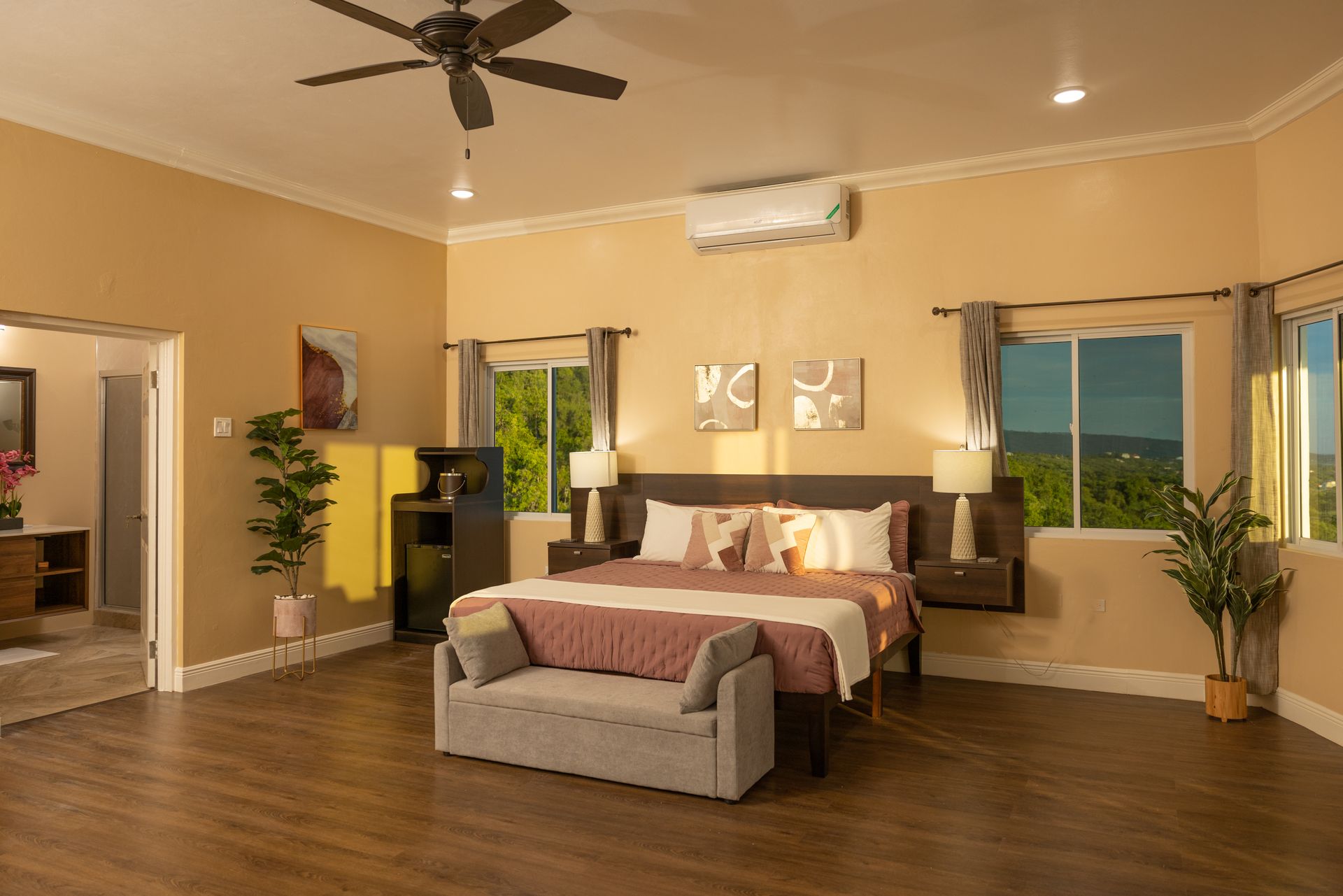 Spacious bedroom with a bed, sofa, windows, and ceiling fan, with a glimpse of an adjacent room.