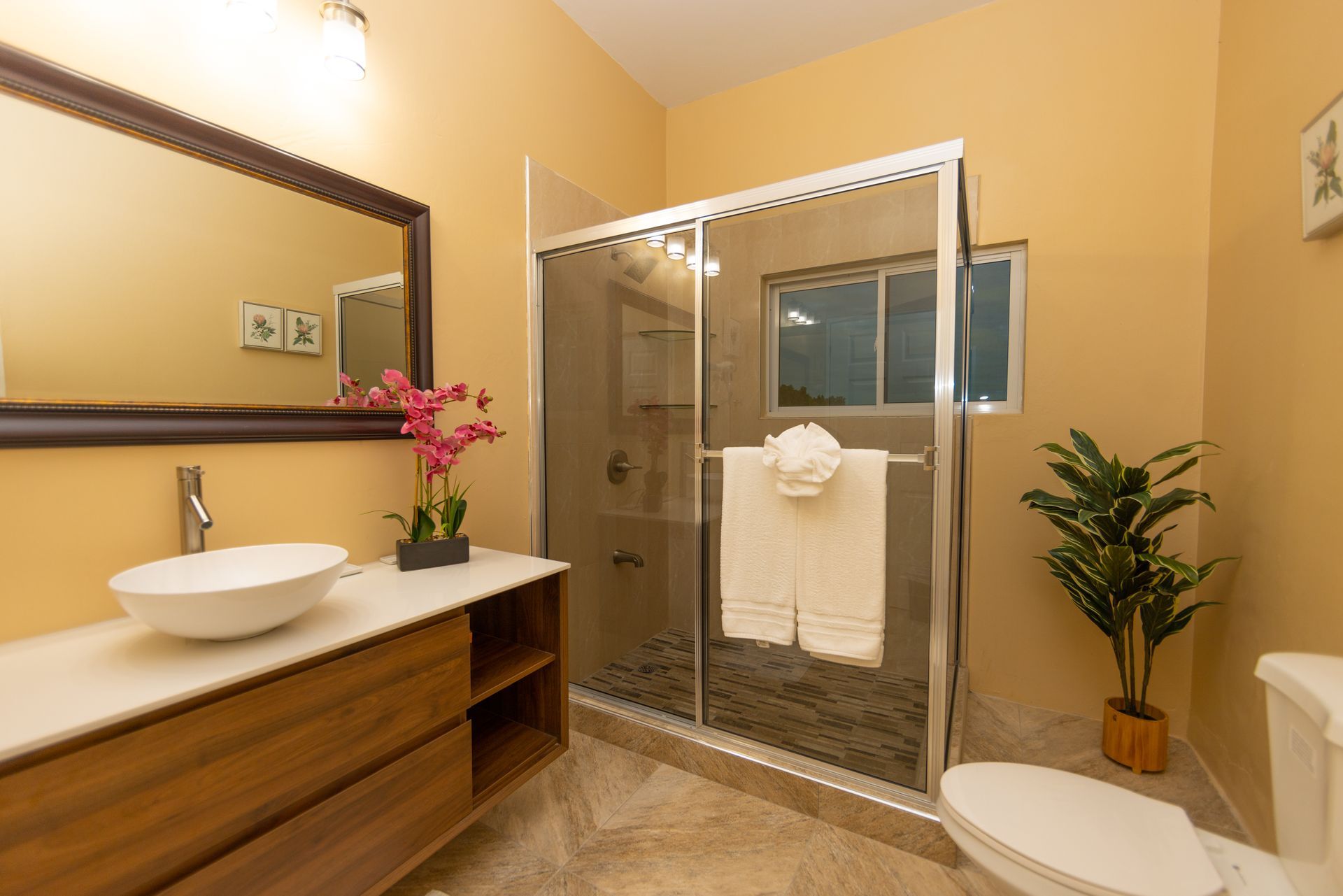 Bathroom with a white sink, shower, and toilet. Yellow walls and wood vanity with a potted plant.
