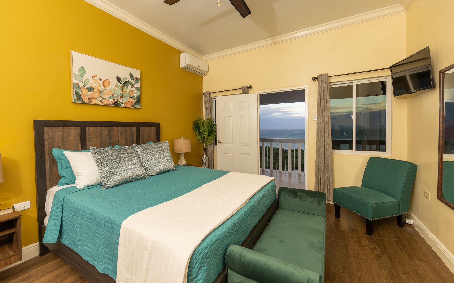 Bedroom with teal bedding, green chair, yellow walls, and a view of the sea.