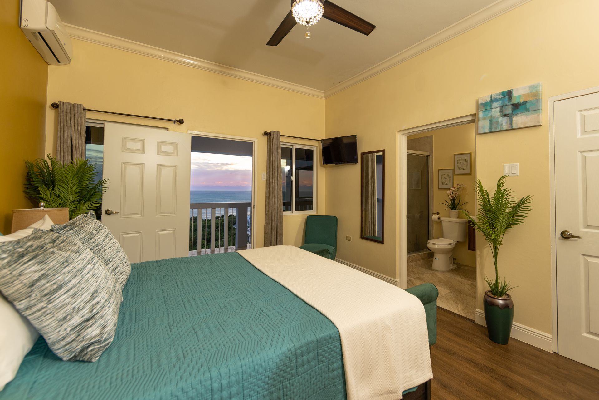 Bedroom with a large bed, teal quilt, and a balcony overlooking the ocean. Light yellow walls and a bathroom visible.