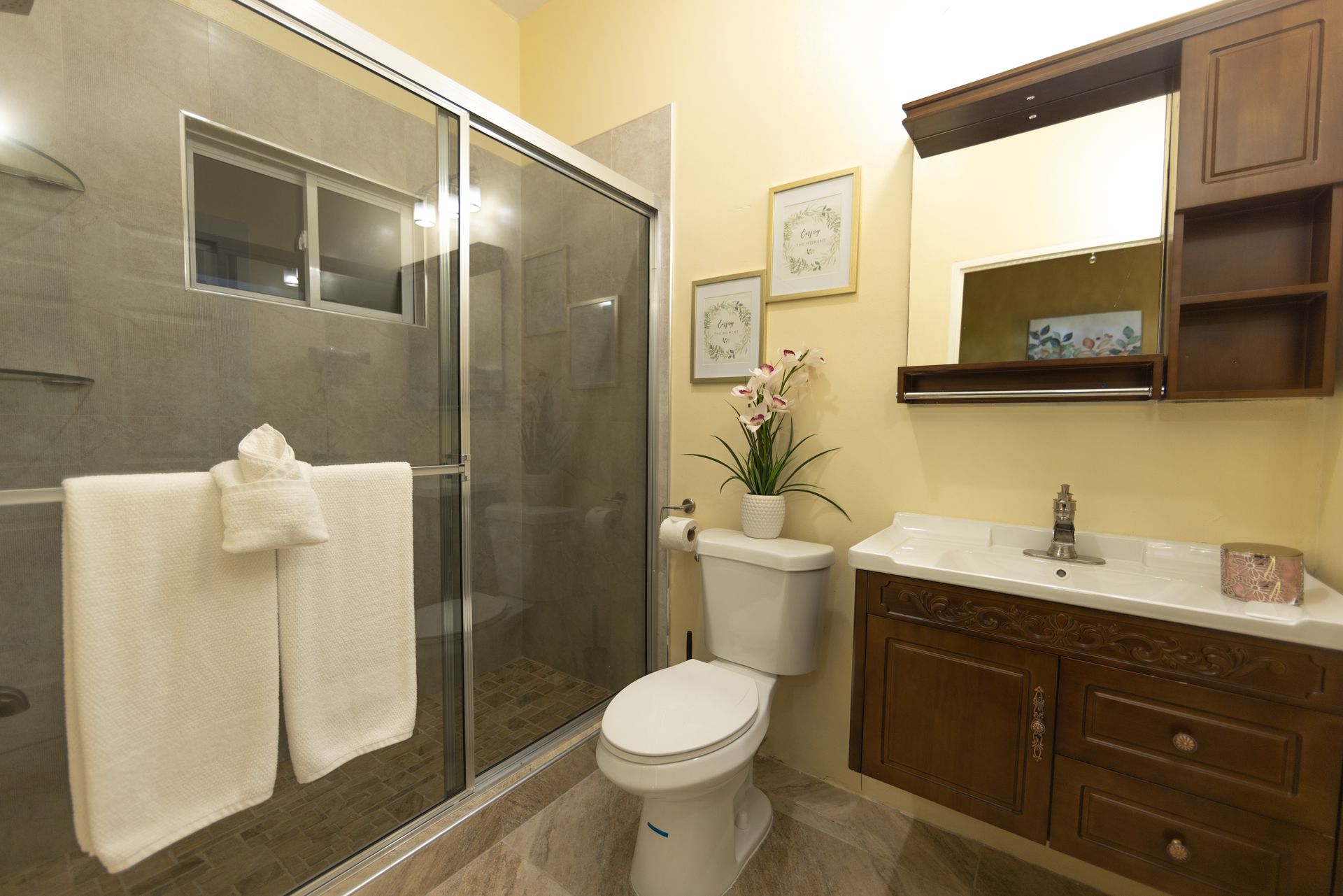 Bathroom with a glass shower, toilet, vanity, and towels. Light yellow walls, brown cabinets.