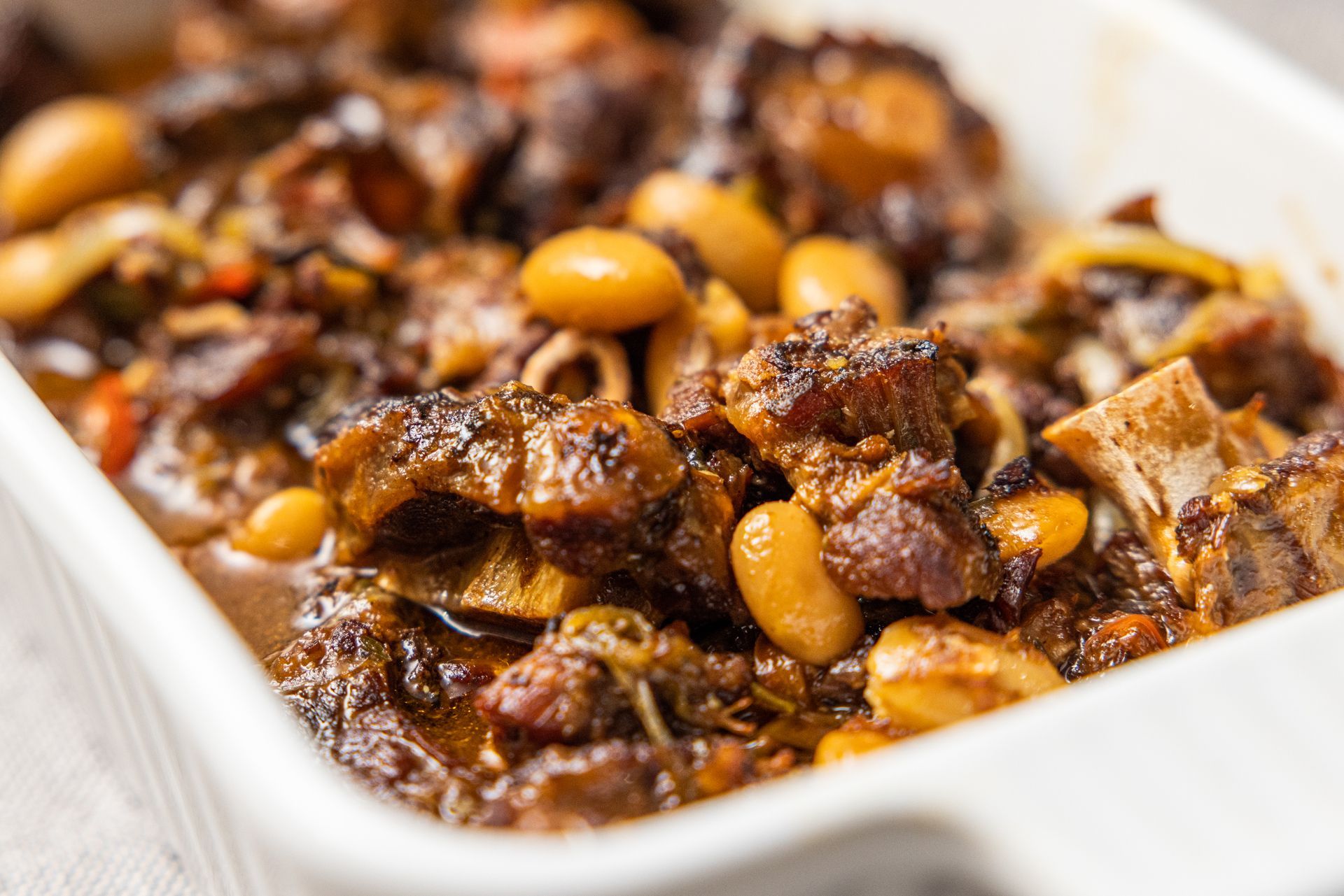Oxtail stew in a white baking dish, with butter beans and vegetables. Brown and saucy.
