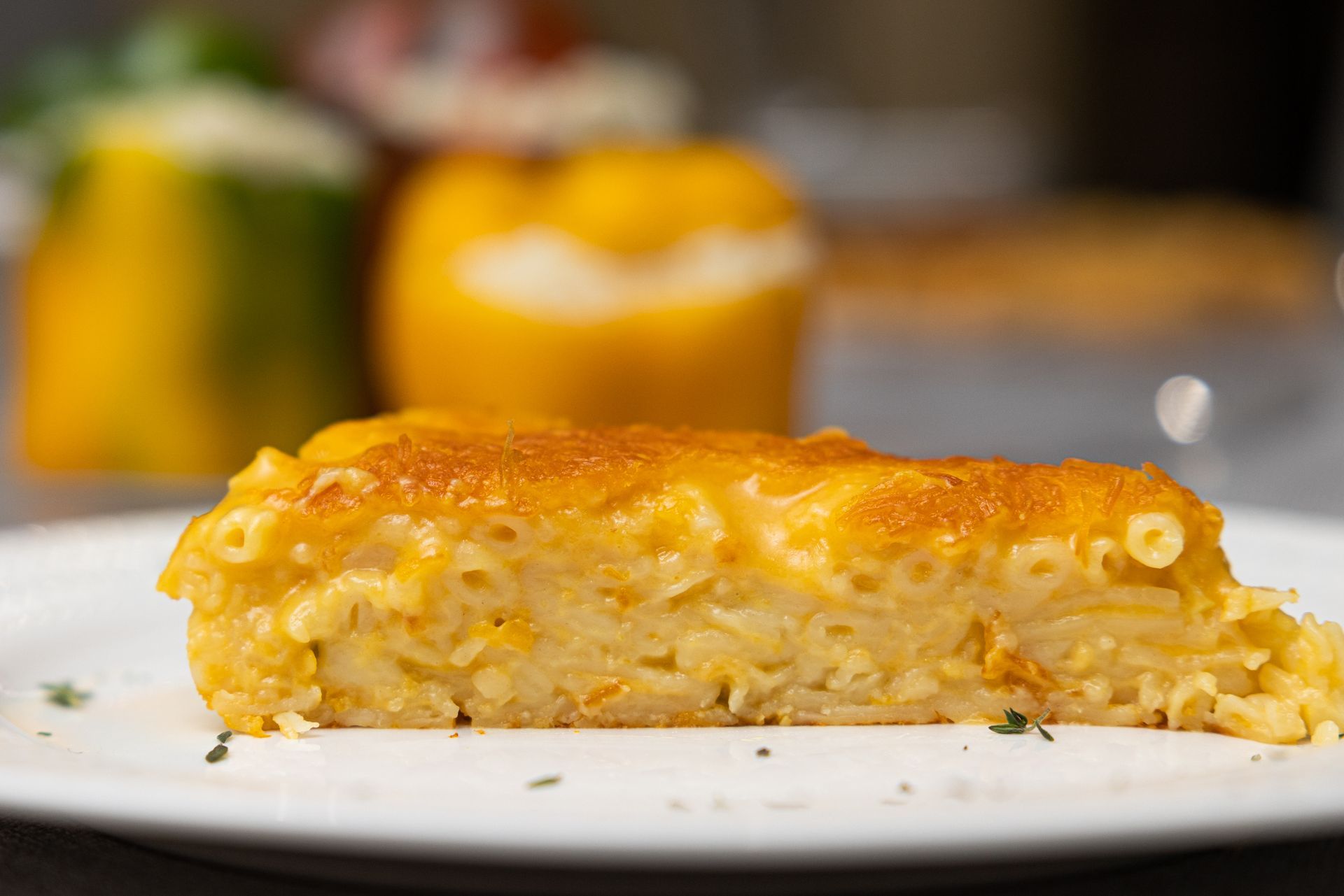 Slice of baked macaroni and cheese on a white plate, with other food items blurred in background.