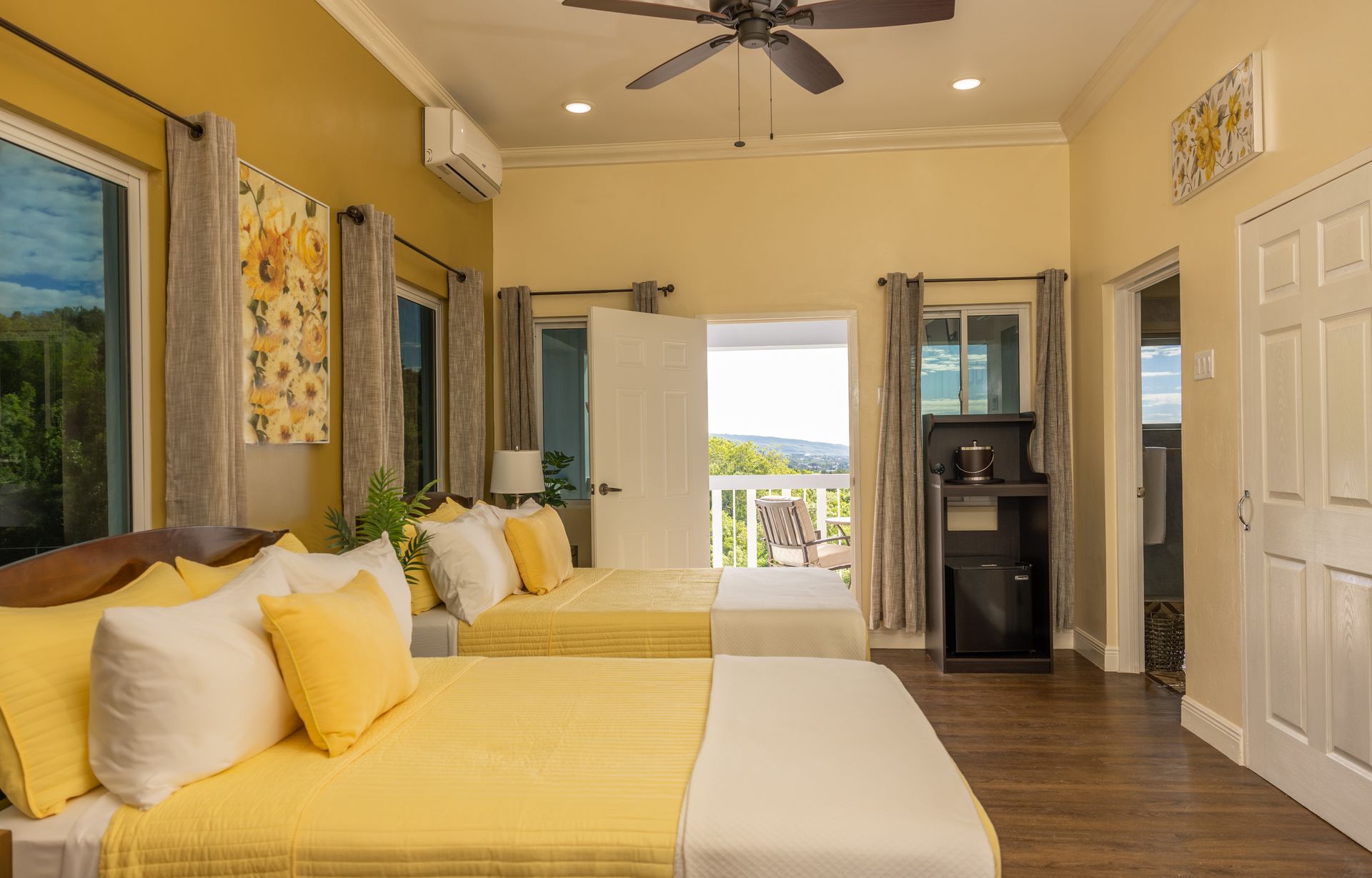 A bright yellow bedroom with two beds, doorway to balcony, and sunflower decor.