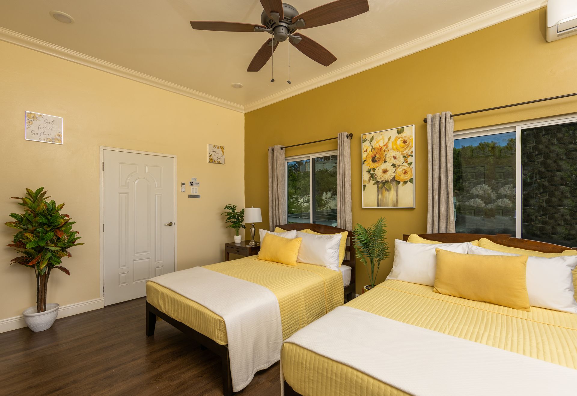 Bedroom with two beds, yellow bedding, floral art, windows, ceiling fan, and hardwood floor.
