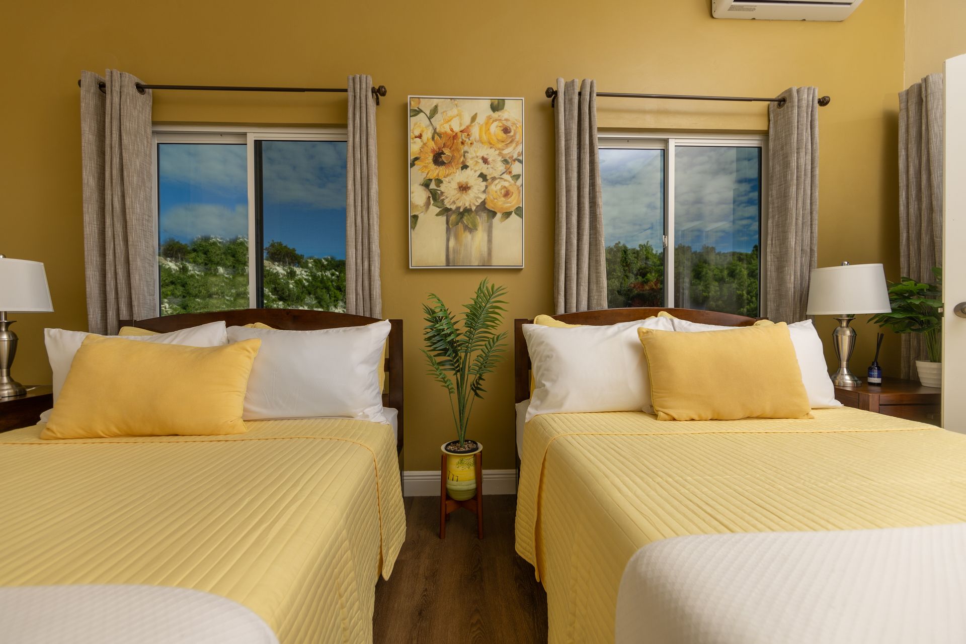 Two beds in a yellow-walled room with floral art between windows showing a blue sky.