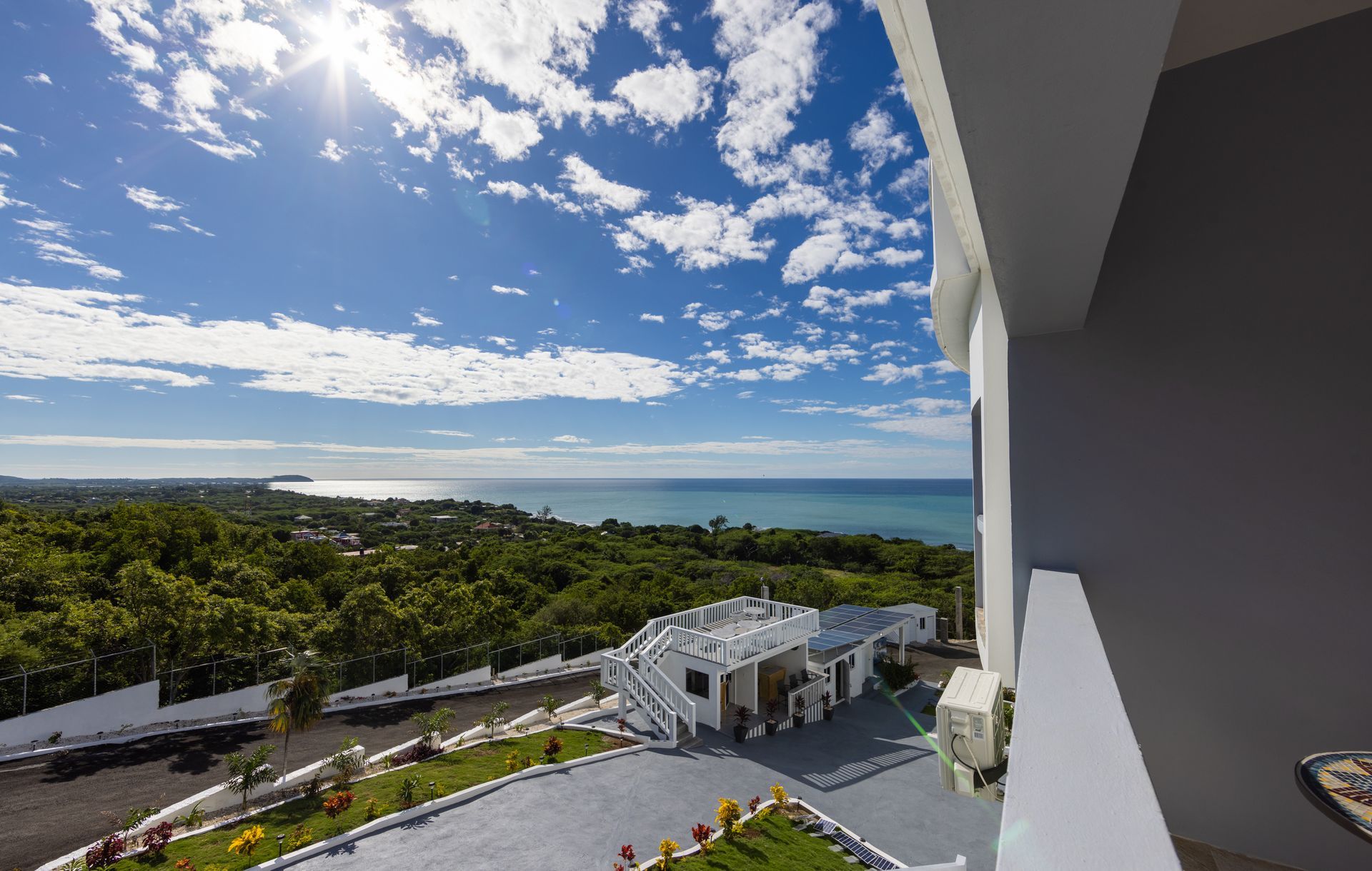 View from a balcony overlooking lush green trees, a road, and the ocean under a bright blue sky with clouds.