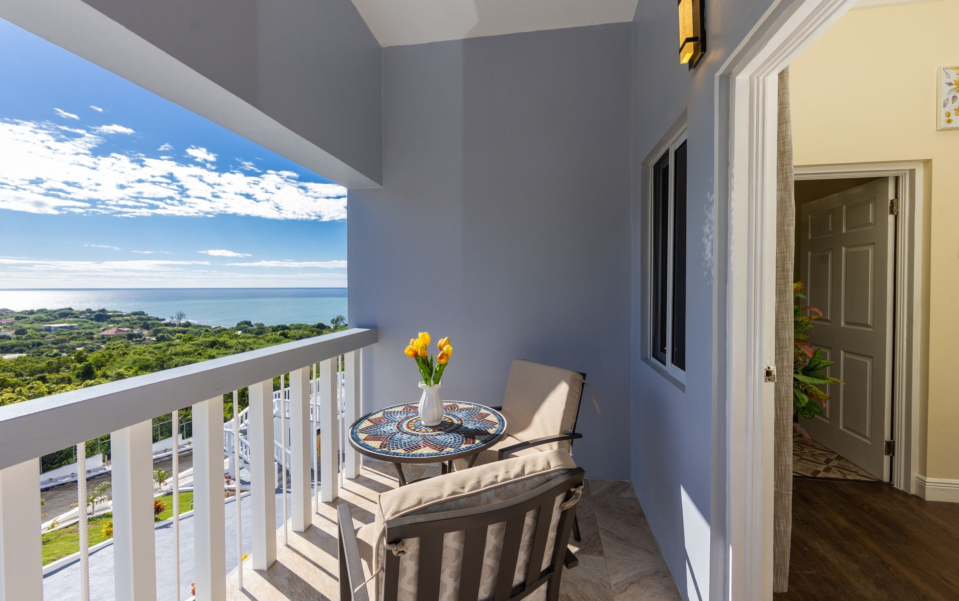 Balcony with ocean view, table, chairs, and flowers. Doorway to a room is visible.