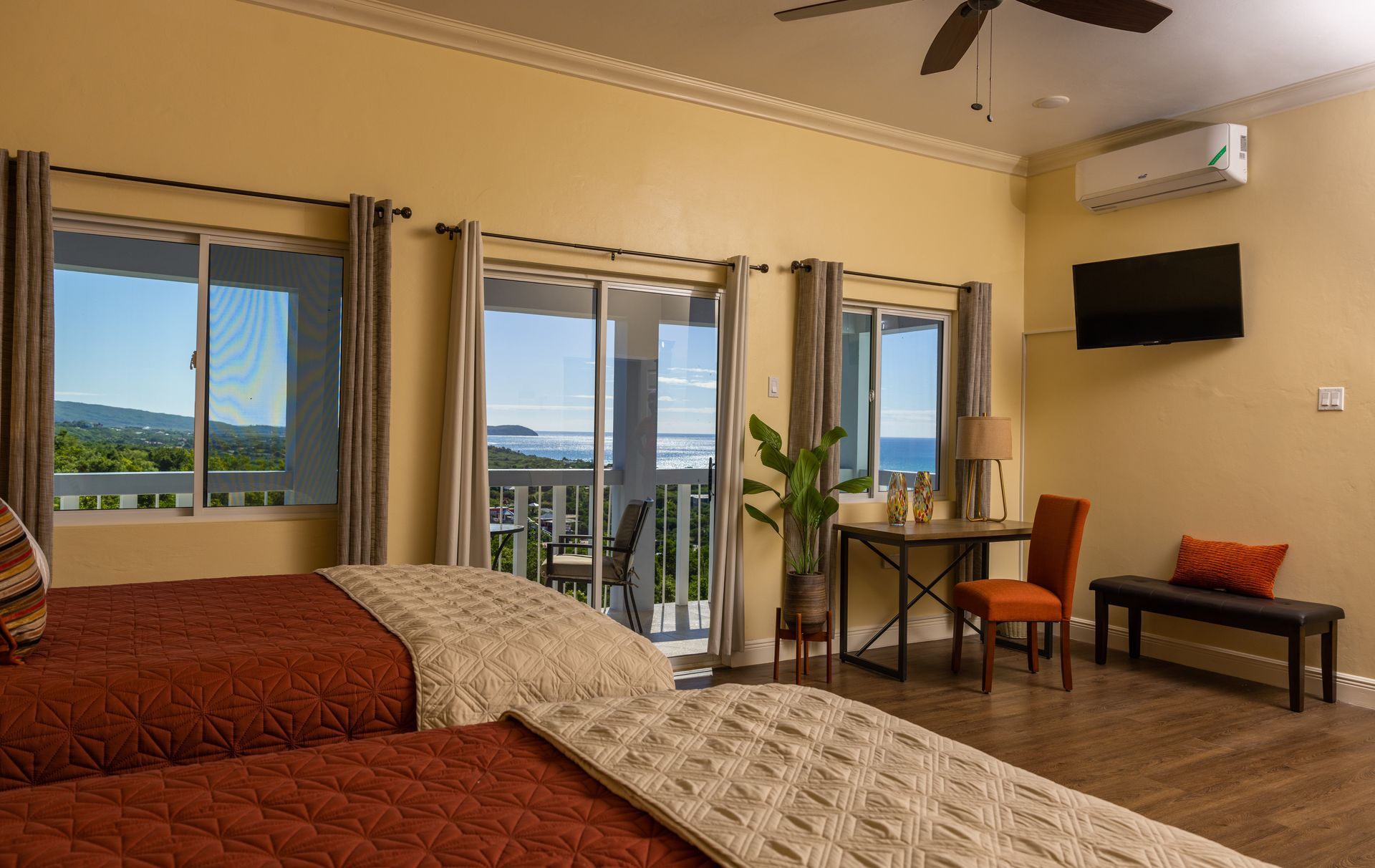 Bedroom with two beds, balcony overlooking ocean, yellow walls, TV, table, and dark curtains.