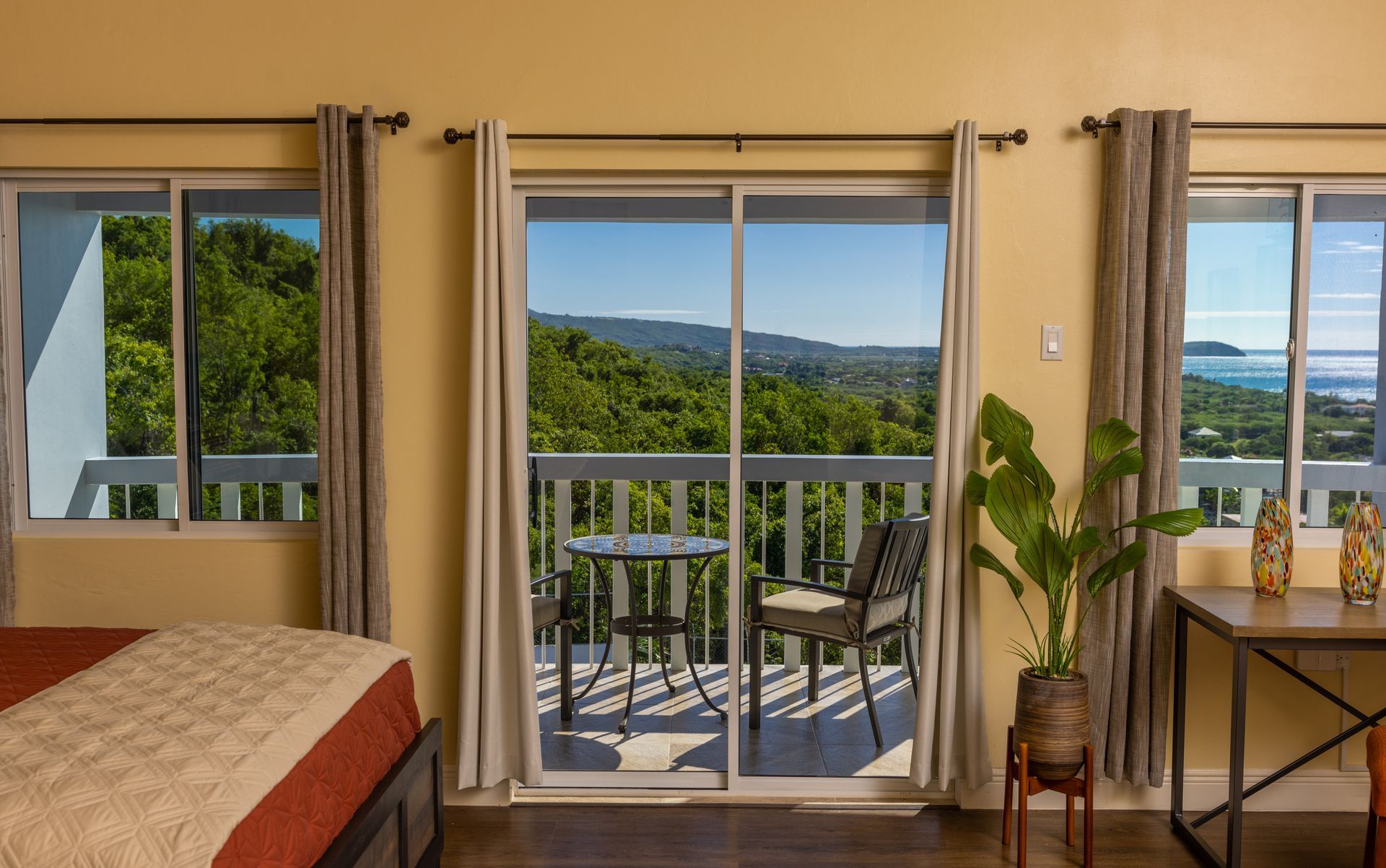 Bedroom with balcony overlooking a lush green landscape and ocean. Furniture includes bed, table, chair, and plant.