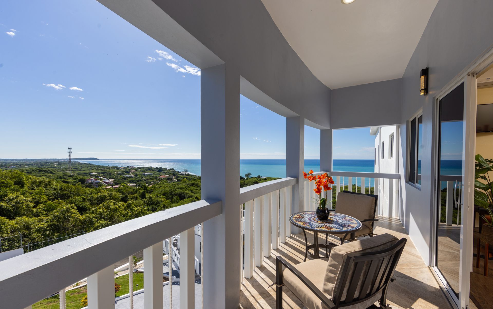 Balcony with two chairs, small table, and ocean view. Bright blue sky, green trees.