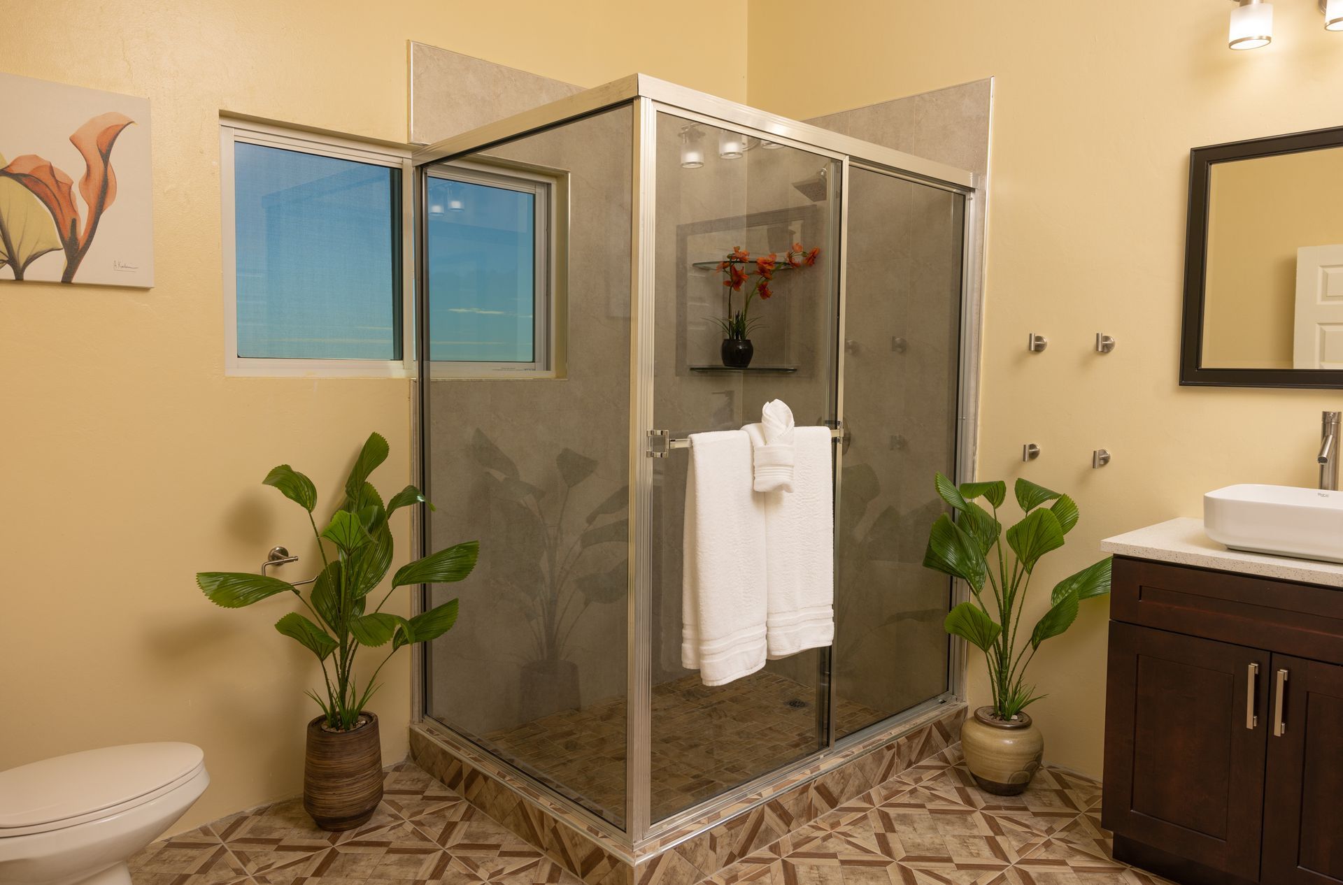 Bathroom with corner shower, vanity, and toilet. Light yellow walls, indoor plants, and a tiled floor.