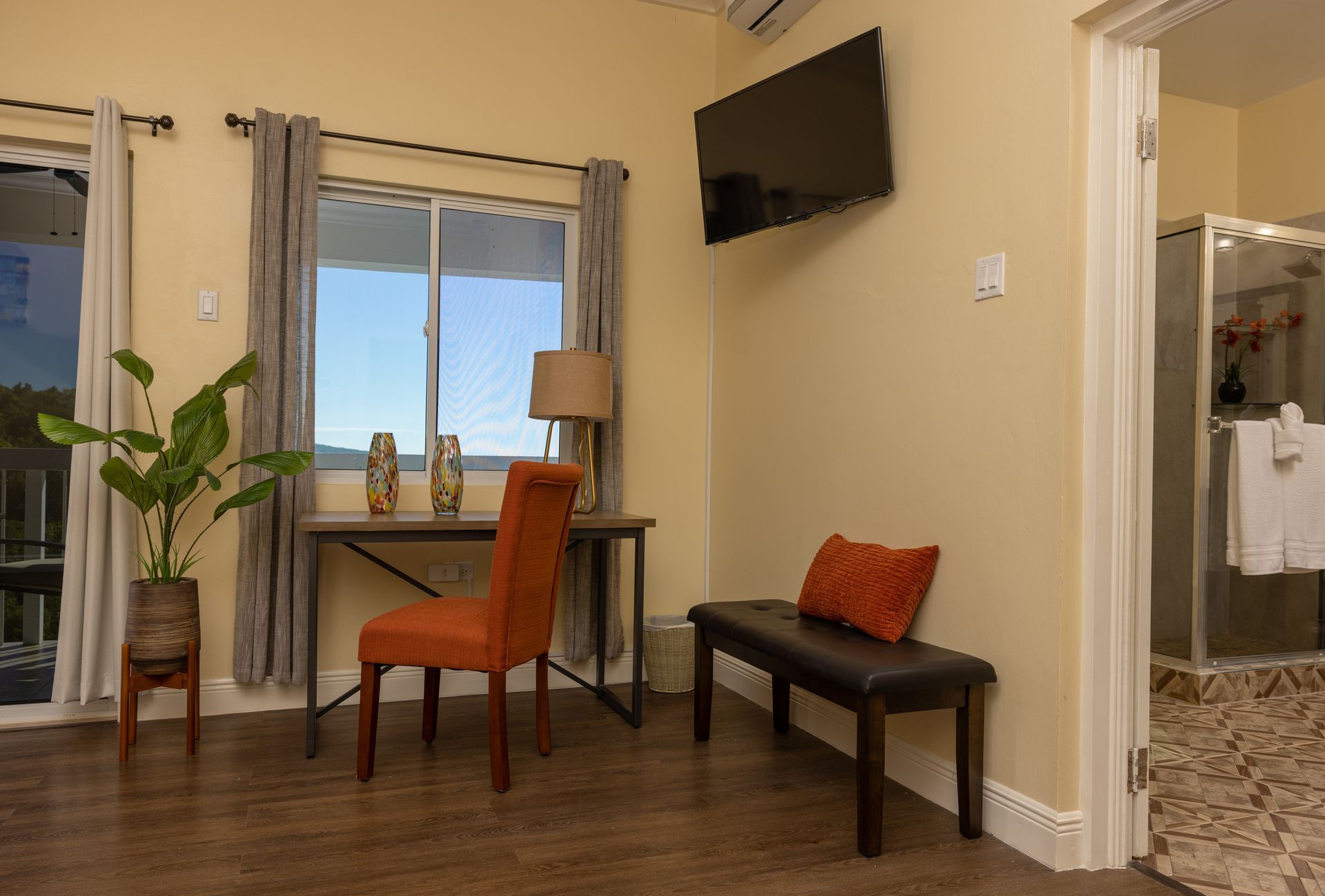 Room with desk, chair, bench, window, TV, and open doorway to a bathroom. Beige walls, wood floors.