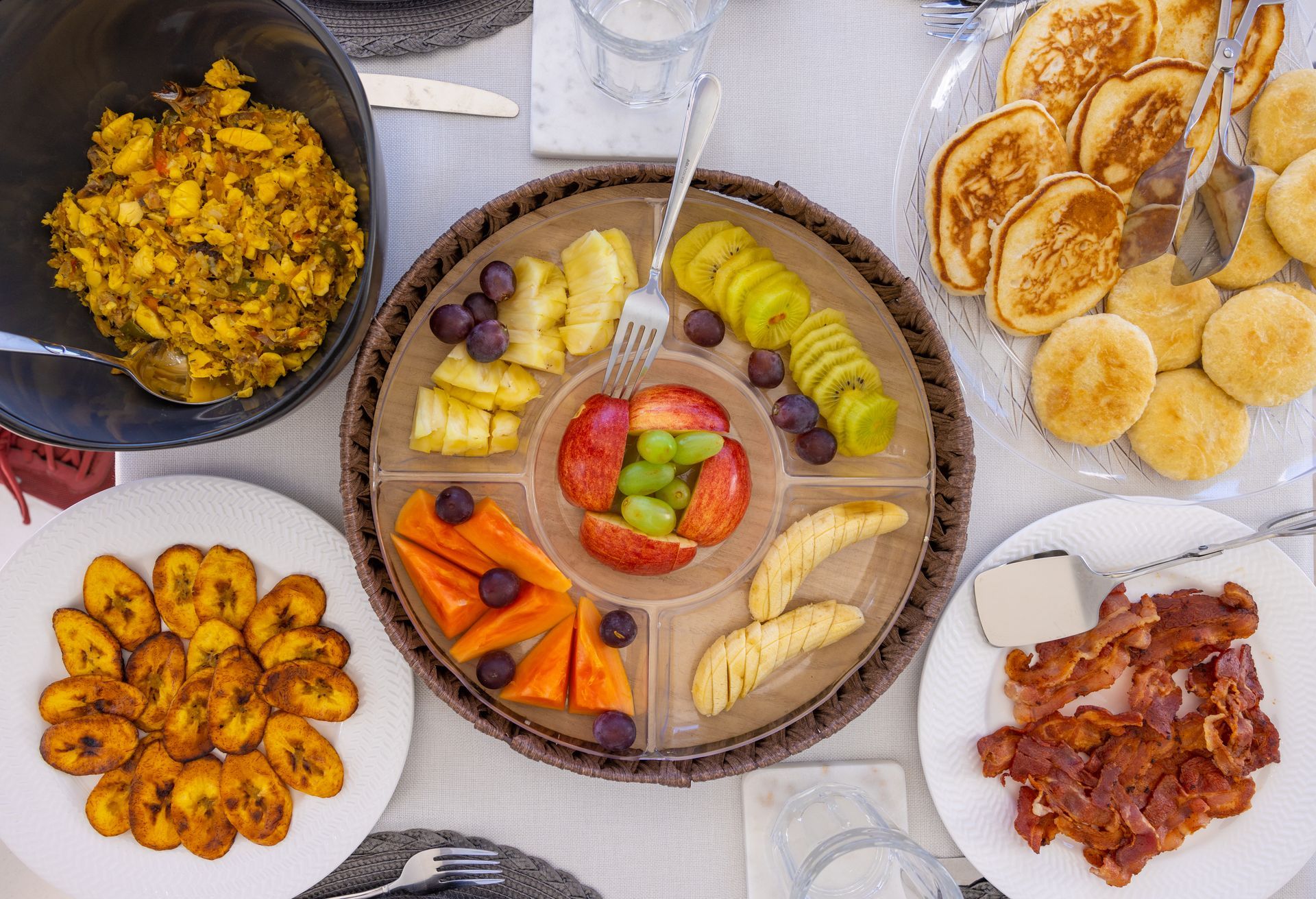 Breakfast spread: Pancakes, bacon, fried plantains, fruit platter with apples, pineapple, and grapes.