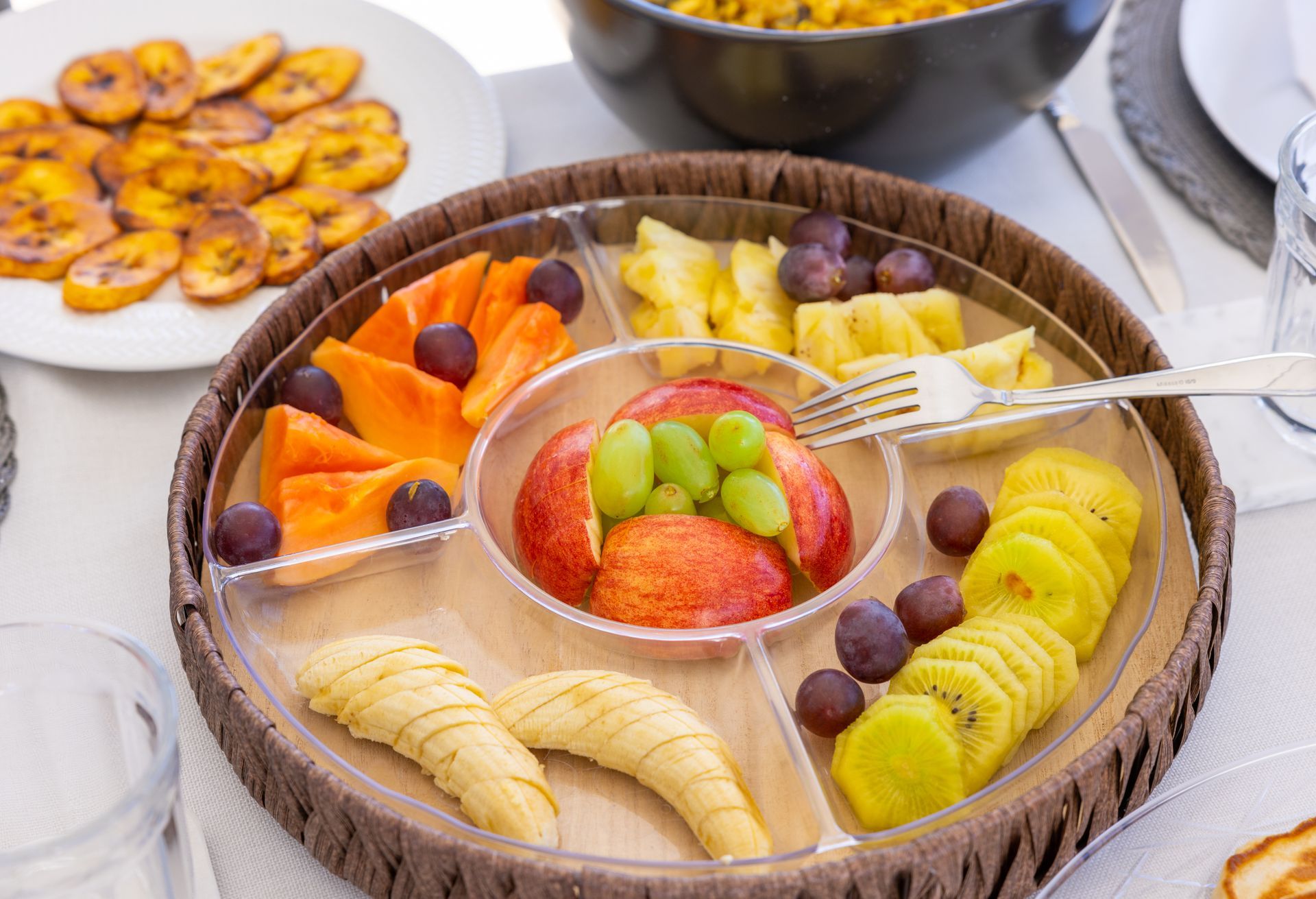 Fruit platter with papaya, pineapple, kiwi, grapes, and apple. Fried plantains in the background.