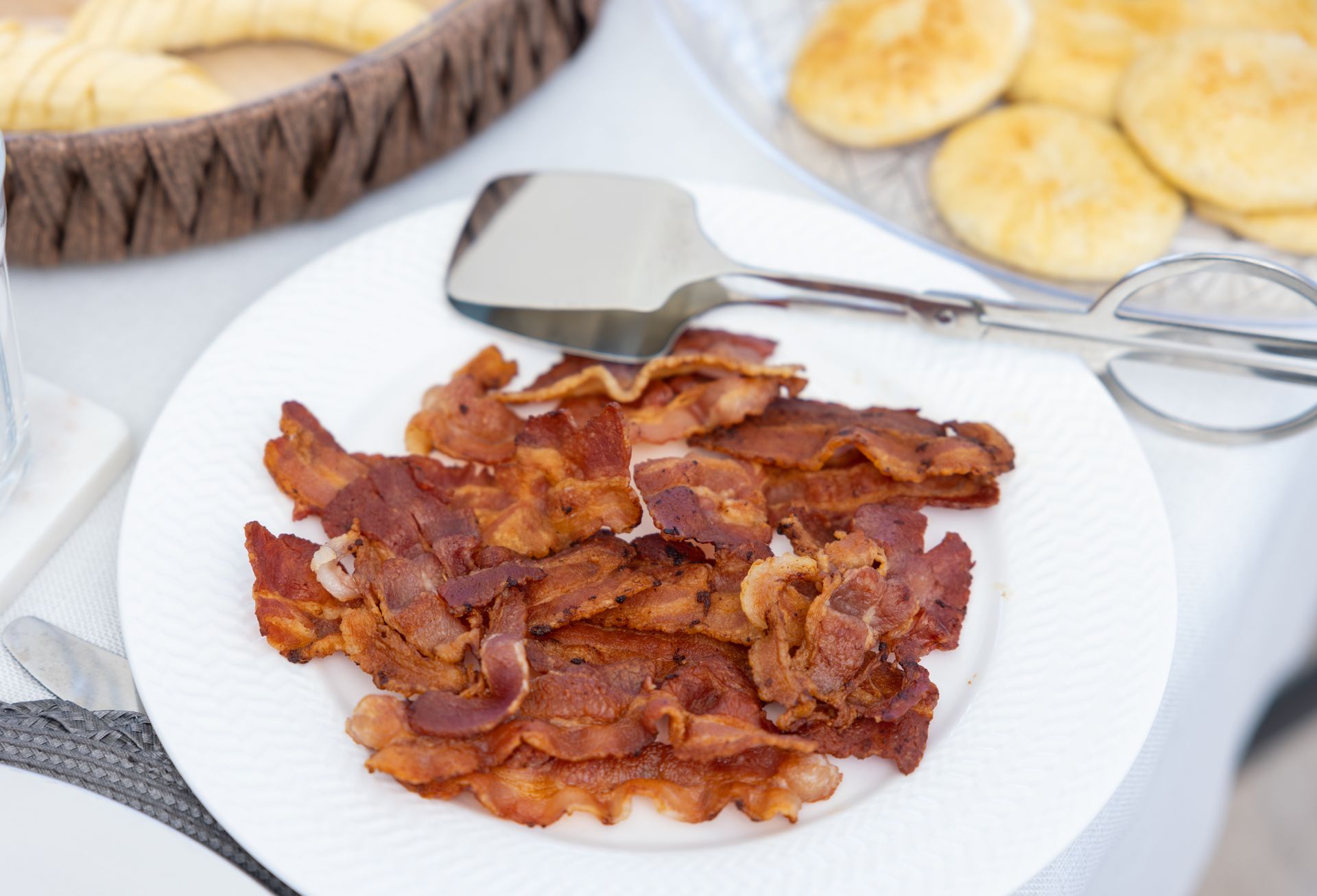 Crispy bacon on a white plate with serving tongs, alongside rolls in a basket.