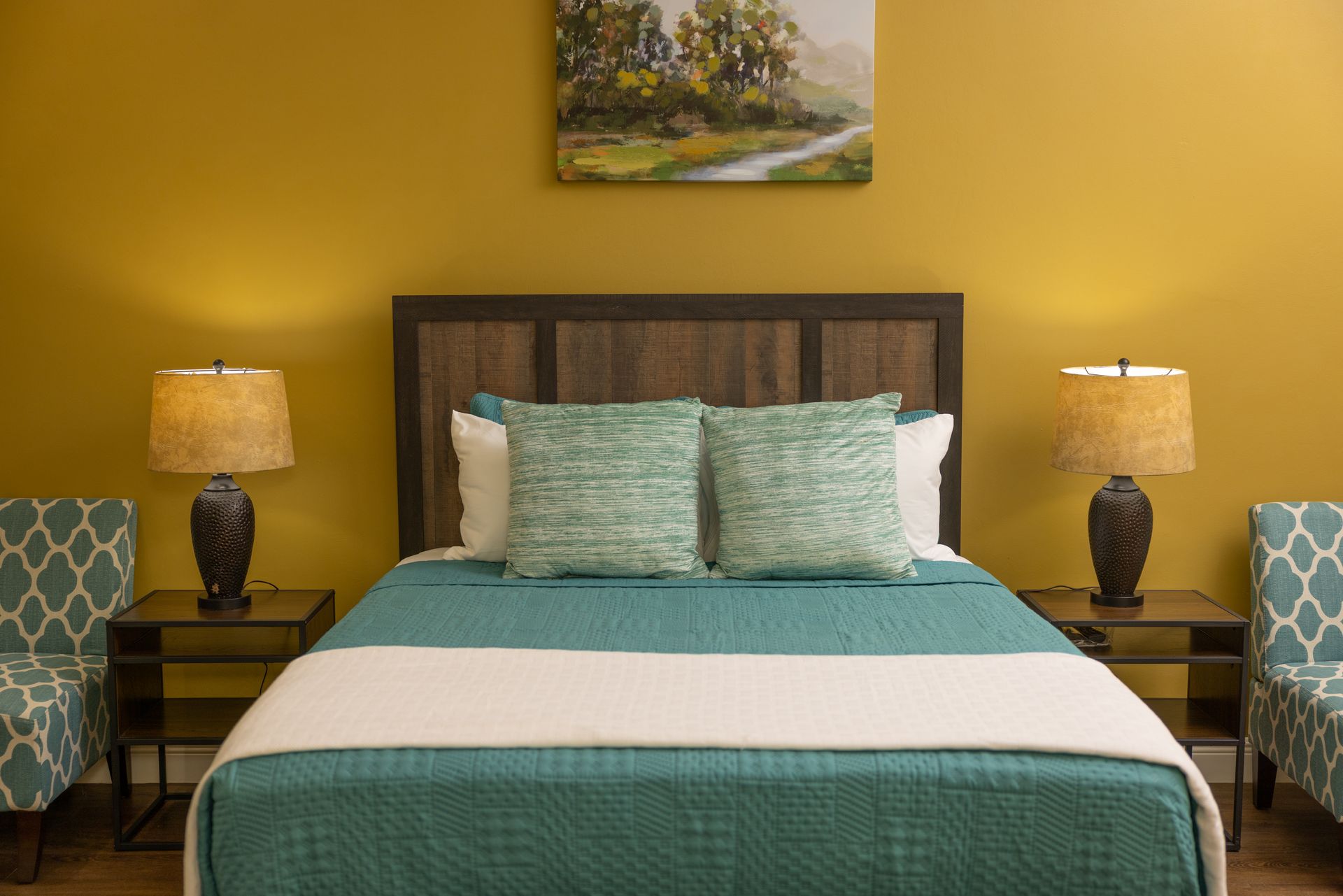 Bedroom with teal bedspread, two matching chairs, bedside lamps, and a painting on the yellow wall.