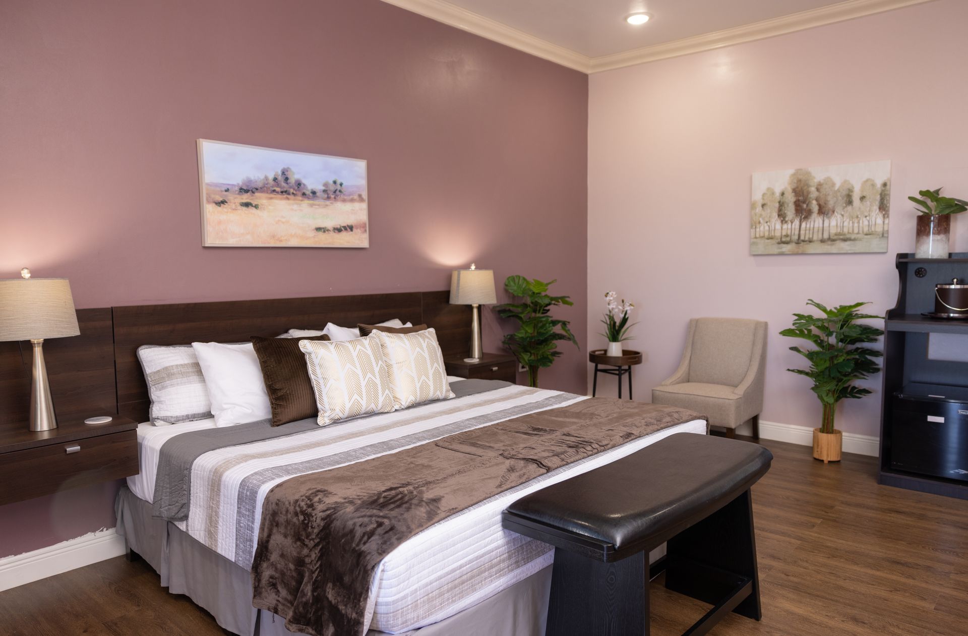 Bedroom with a bed, bench, side tables, artwork, plants, and a small refrigerator. Walls are mauve and beige.