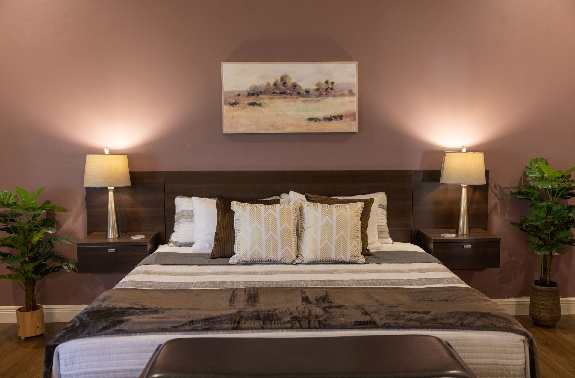 Bedroom with queen bed, brown and white bedding, two lamps, floating nightstands, artwork, and plants against a purple wall.