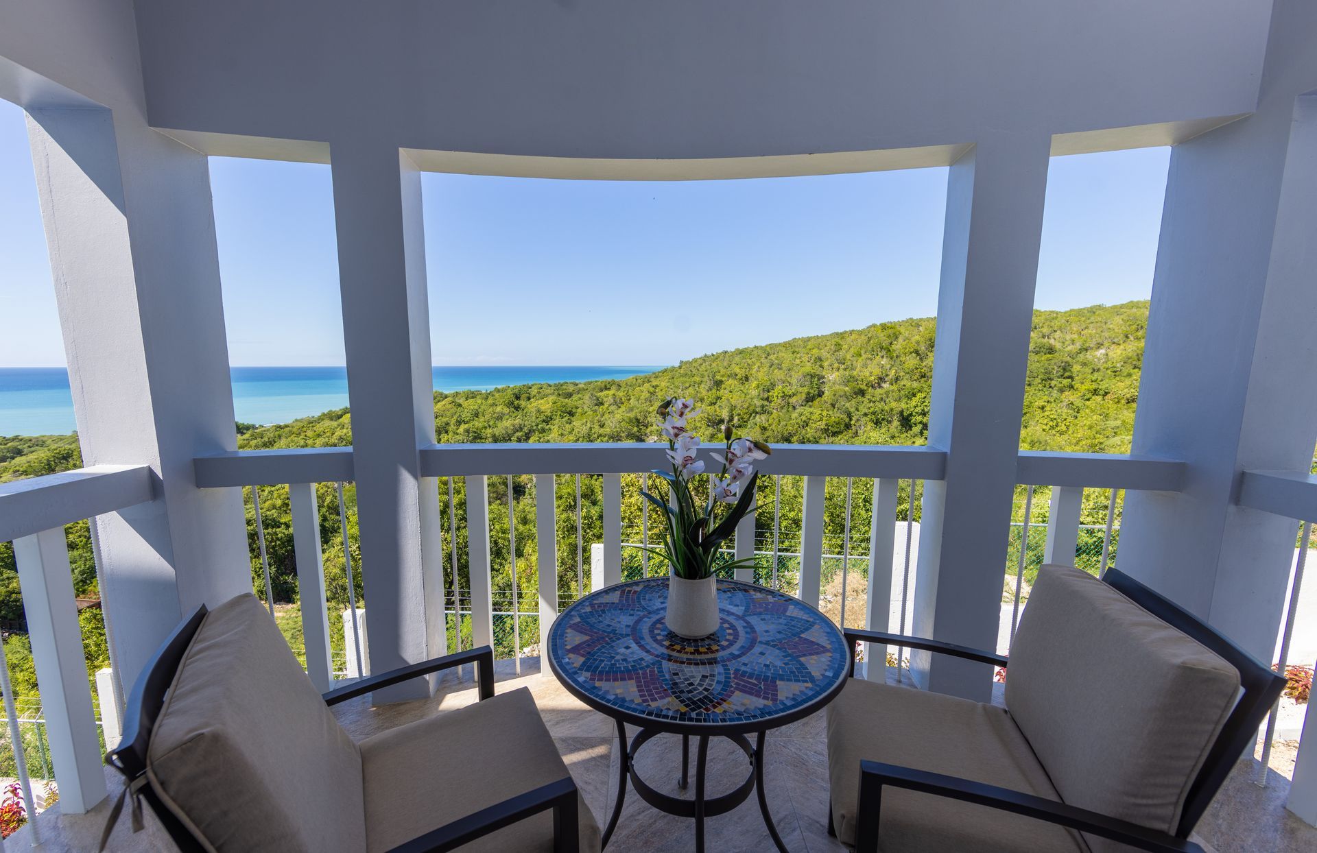 Balcony with two chairs, small table, and vase of flowers, overlooking lush green forest and ocean.