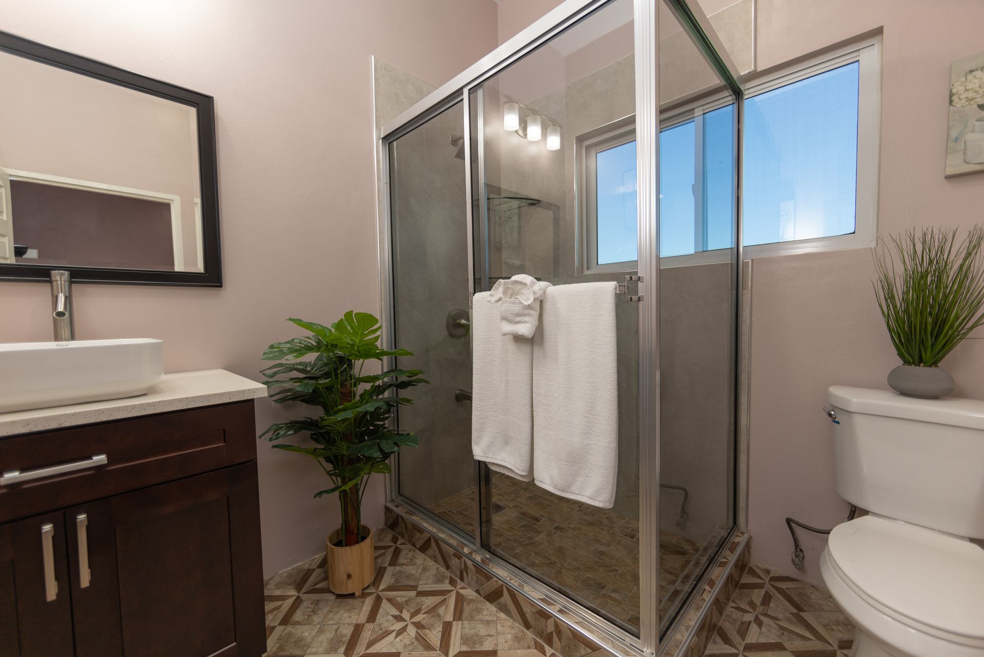 Bathroom with shower, toilet, vanity, and decorative plant against light pink walls; window with blue sky.